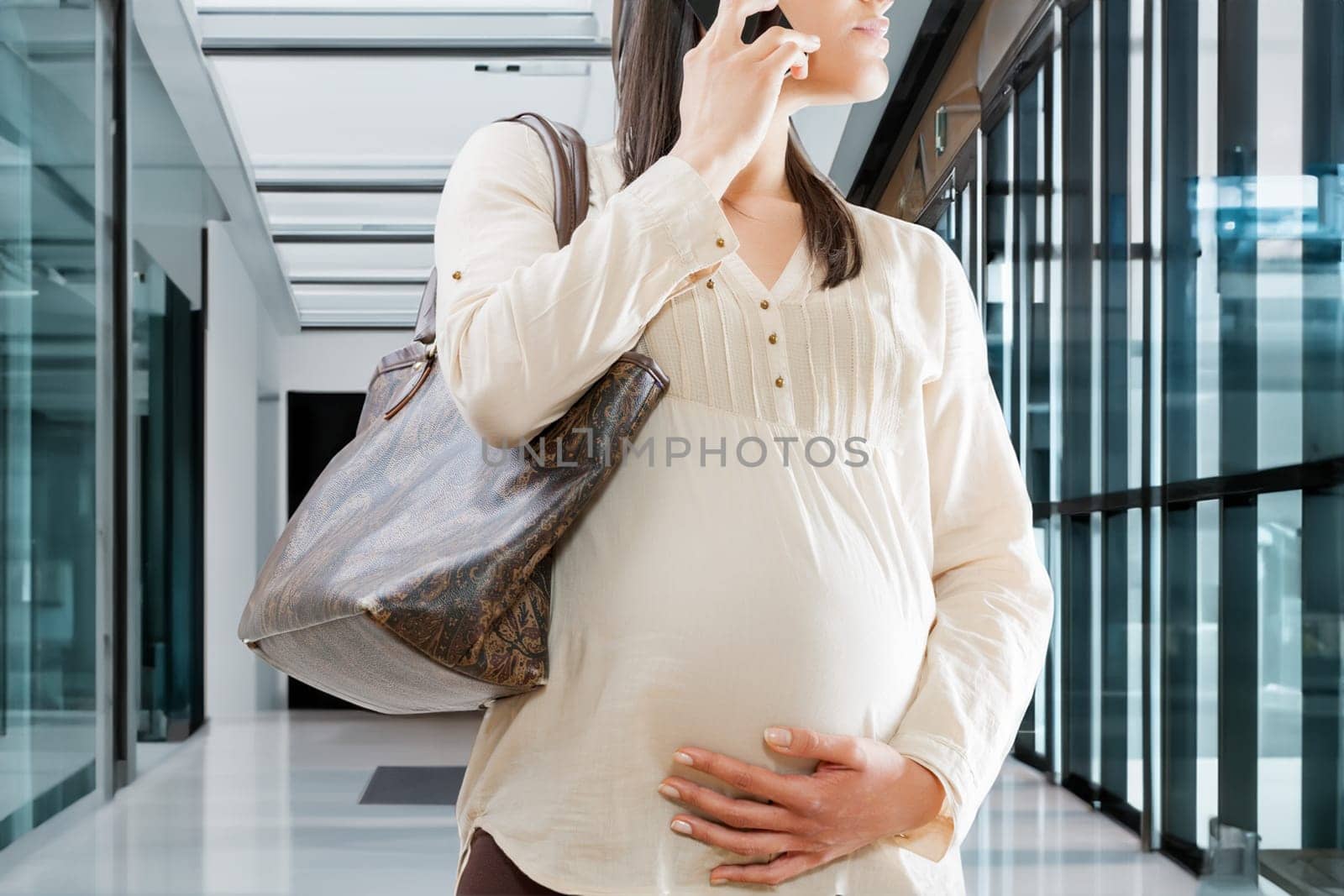 unrecognizable pregnant woman is engrossed in a phone conversation in a corporate environment. Working late into pregnancy, out of necessity or personal commitment and conviction