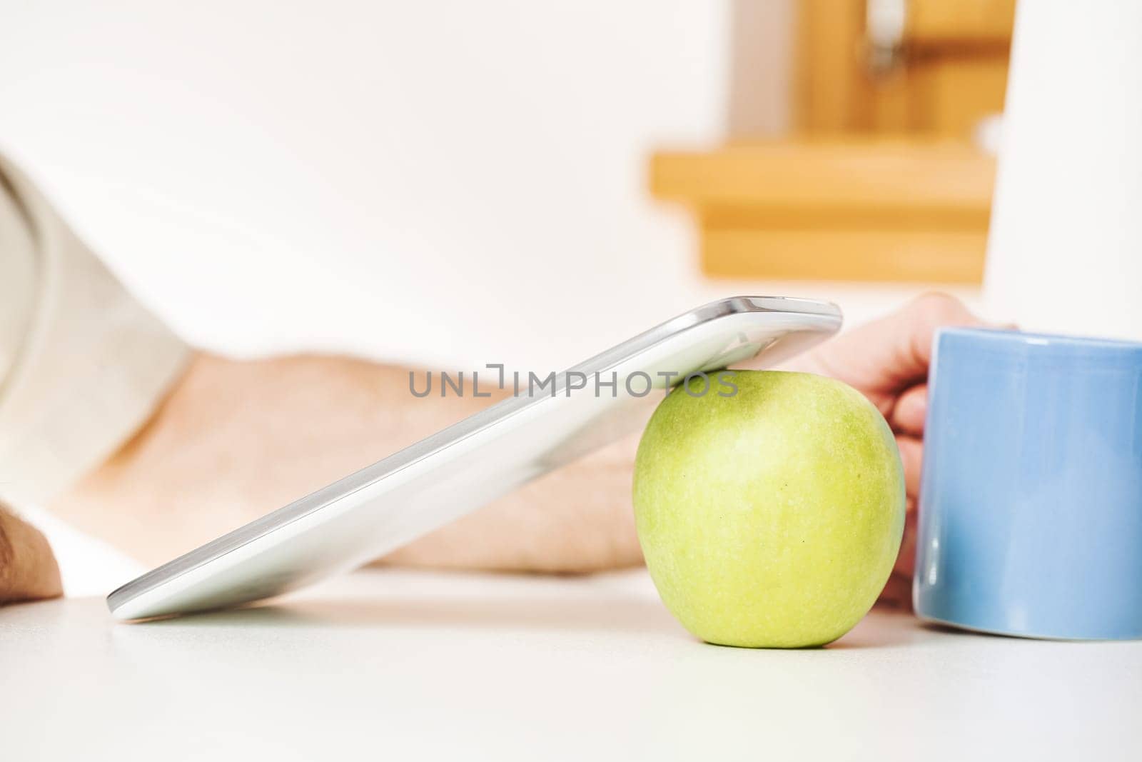 In a symbolic setup, a digital tablet stands on a green apple, accompanied by a blue mug in a kitchen. The presence of a man's arm, face not visible, indicates a motive: to utilize these tools to bene