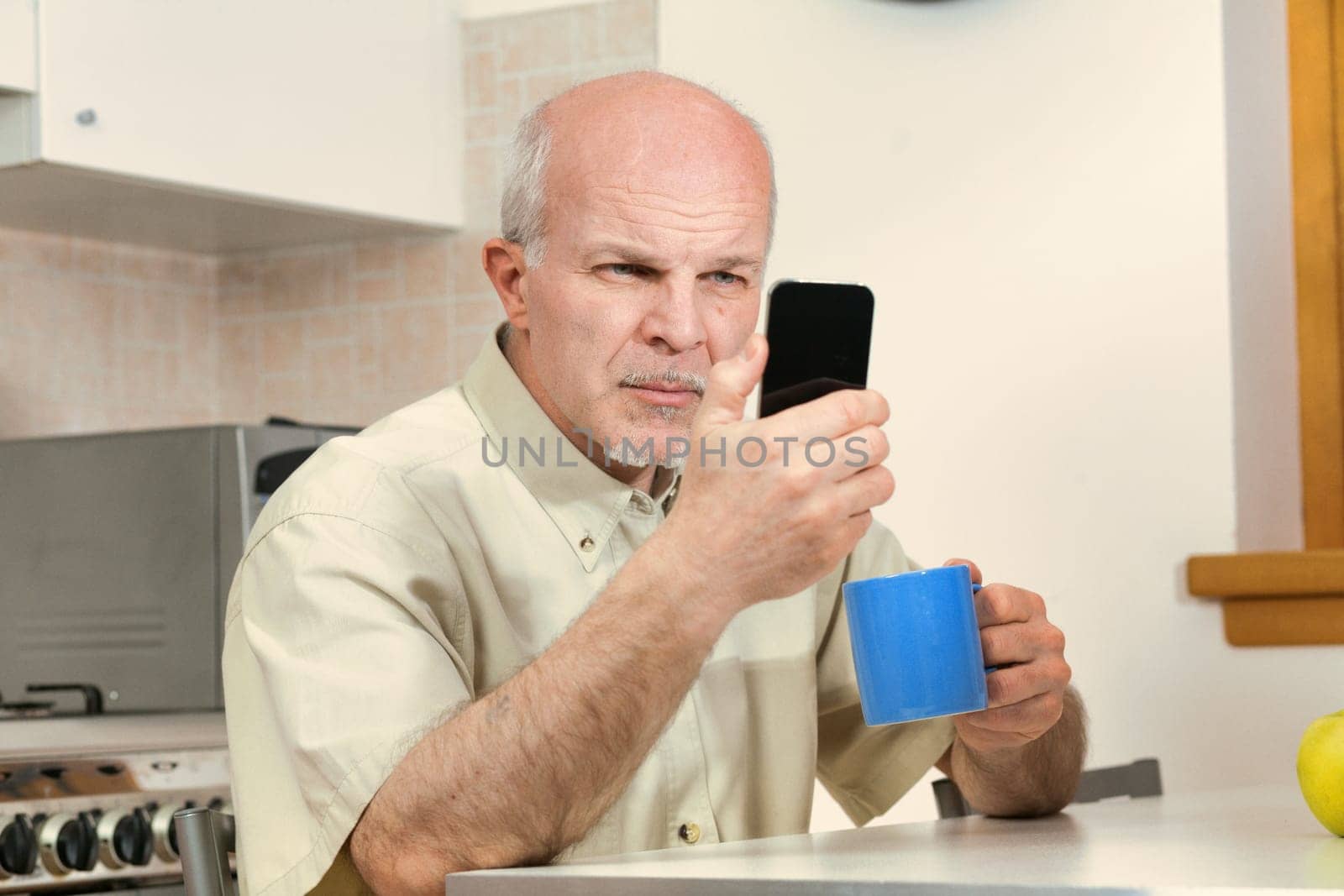 Senior man in kitchen squints at his smartphone during breakfast. Today's phone designs may neglect the visually impaired. A visit to the optometrist and glasses could aid him