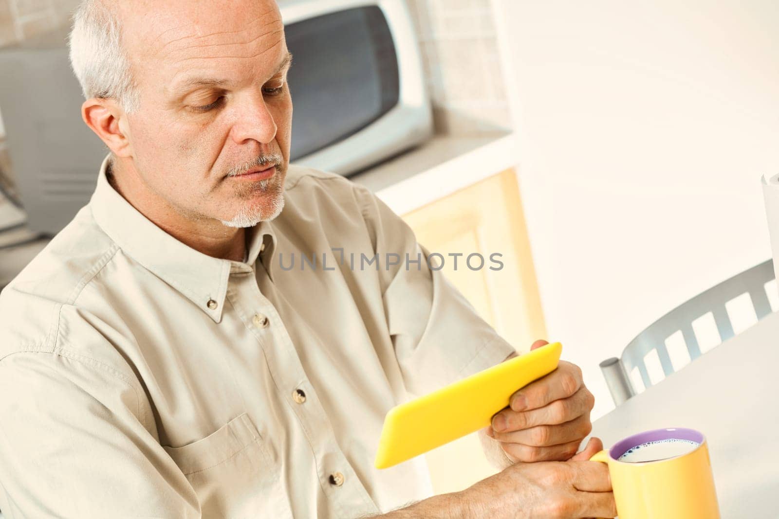 In his kitchen, a senior finds using his smartphone challenging. He squints over breakfast, indicative of phones not designed for vision issues. Perhaps glasses could assist him