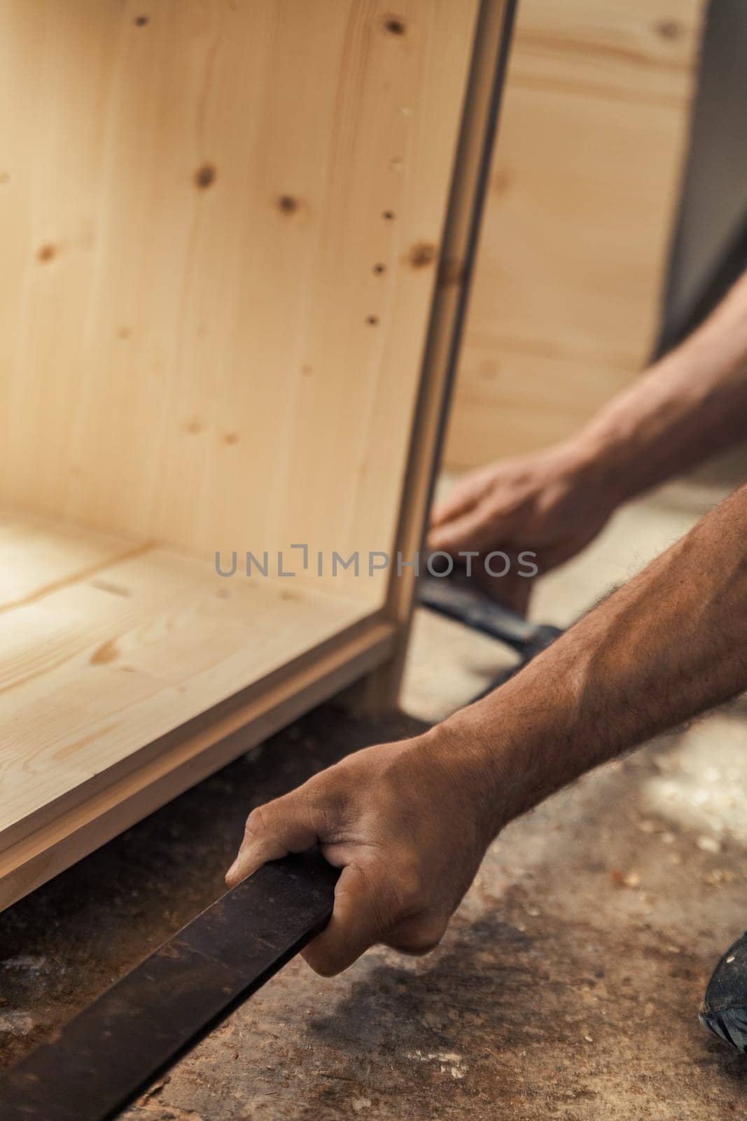 The strong hand of a carpenter firmly grasps a metal tool to assist with a heavy-duty job in the construction of furniture. Warm sunlight streams in through the windows and bathes the surfaces of the