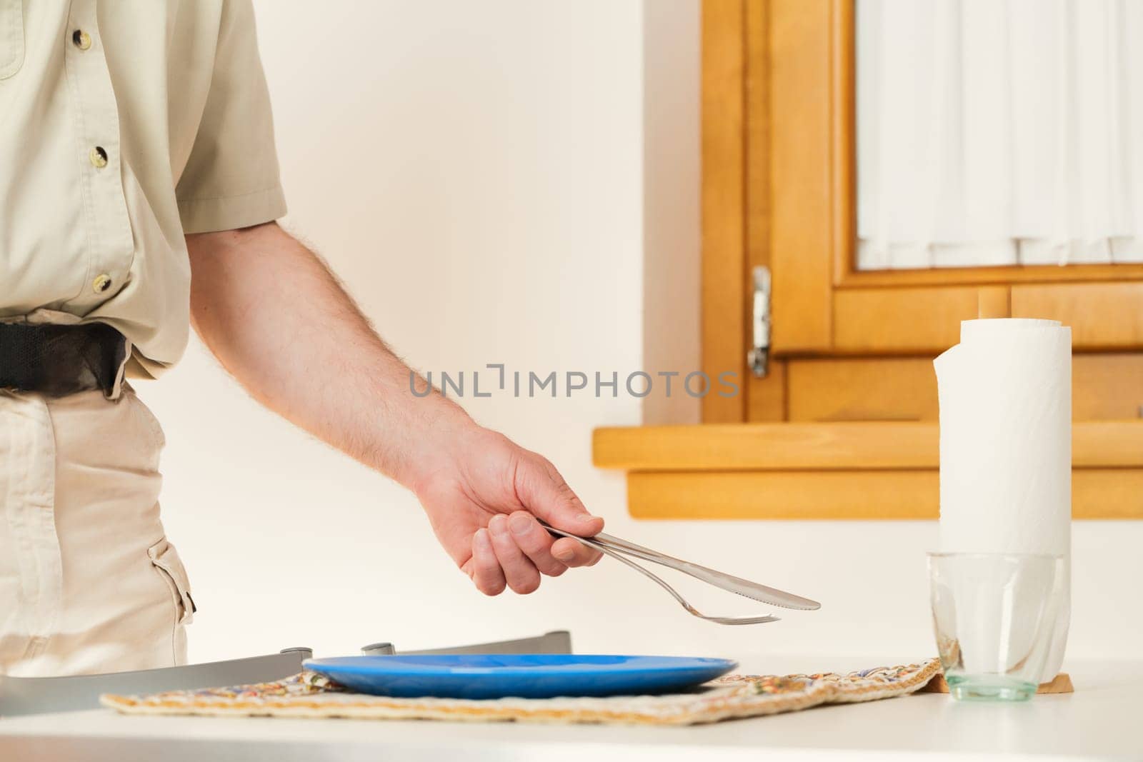 In the intimate setting of his kitchen, an unidentified man's arm reaches out. He's setting the table, laying a knife and fork adjacent to the plate. His neat attire implies a solitary existence