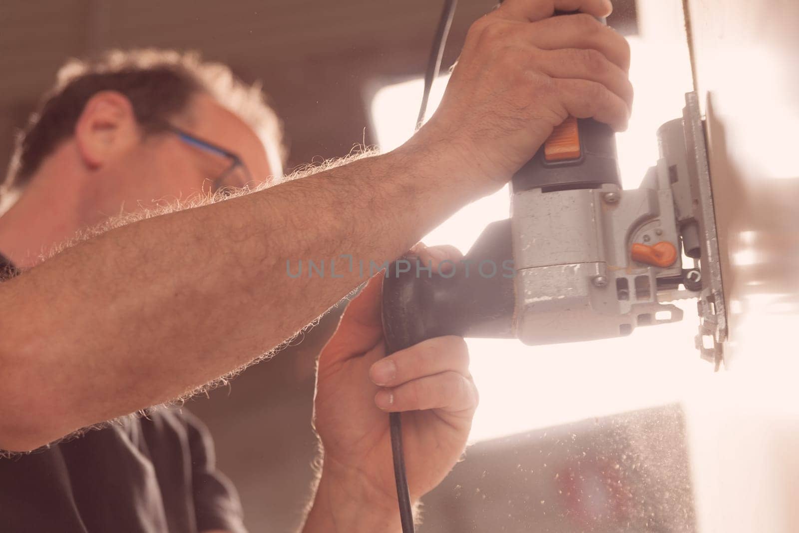close-up on craftsman's hand using a sanding or cutting tool that produces chips and sawdust visible against the light, he is out of focus in the background