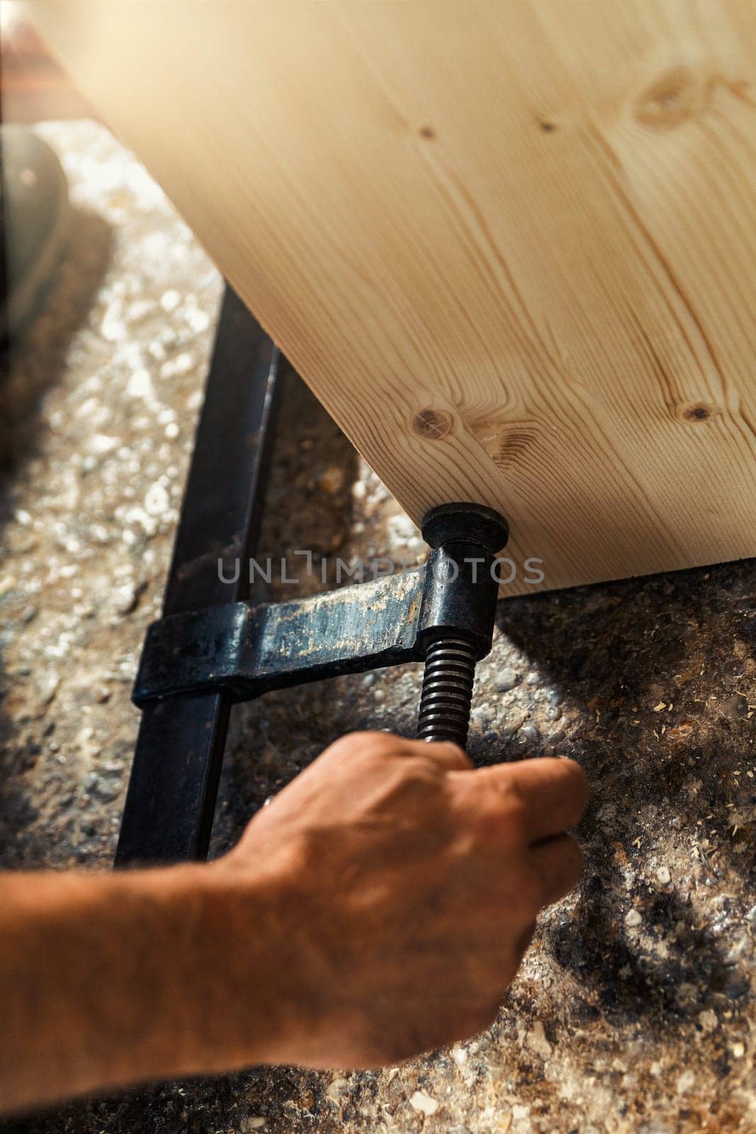 The carpenter's hand secures two parts of a furniture piece with a wooden screw clamp in his artisanal workshop, illuminated by warm sunlight pouring through the window