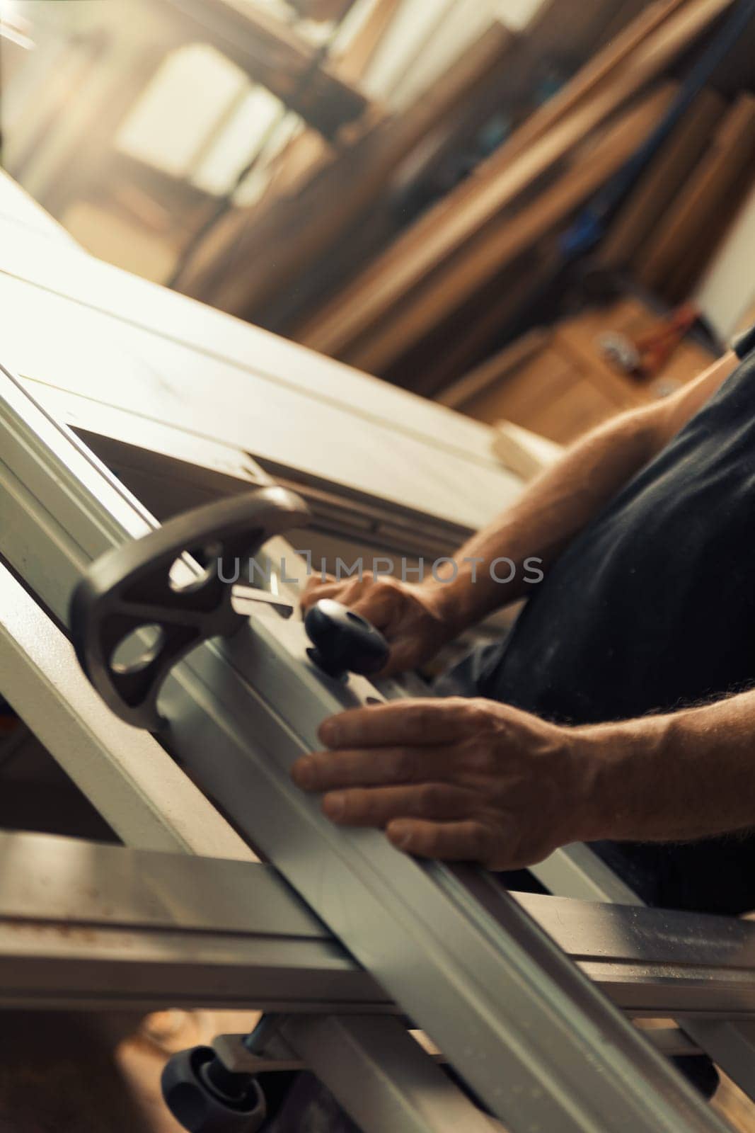 n a woodworking shop, a man is positioning a wooden shelf or board onto a lumber processing machine. Strong male hands and large amounts of wood are visible, as well as the metal structures formed by
