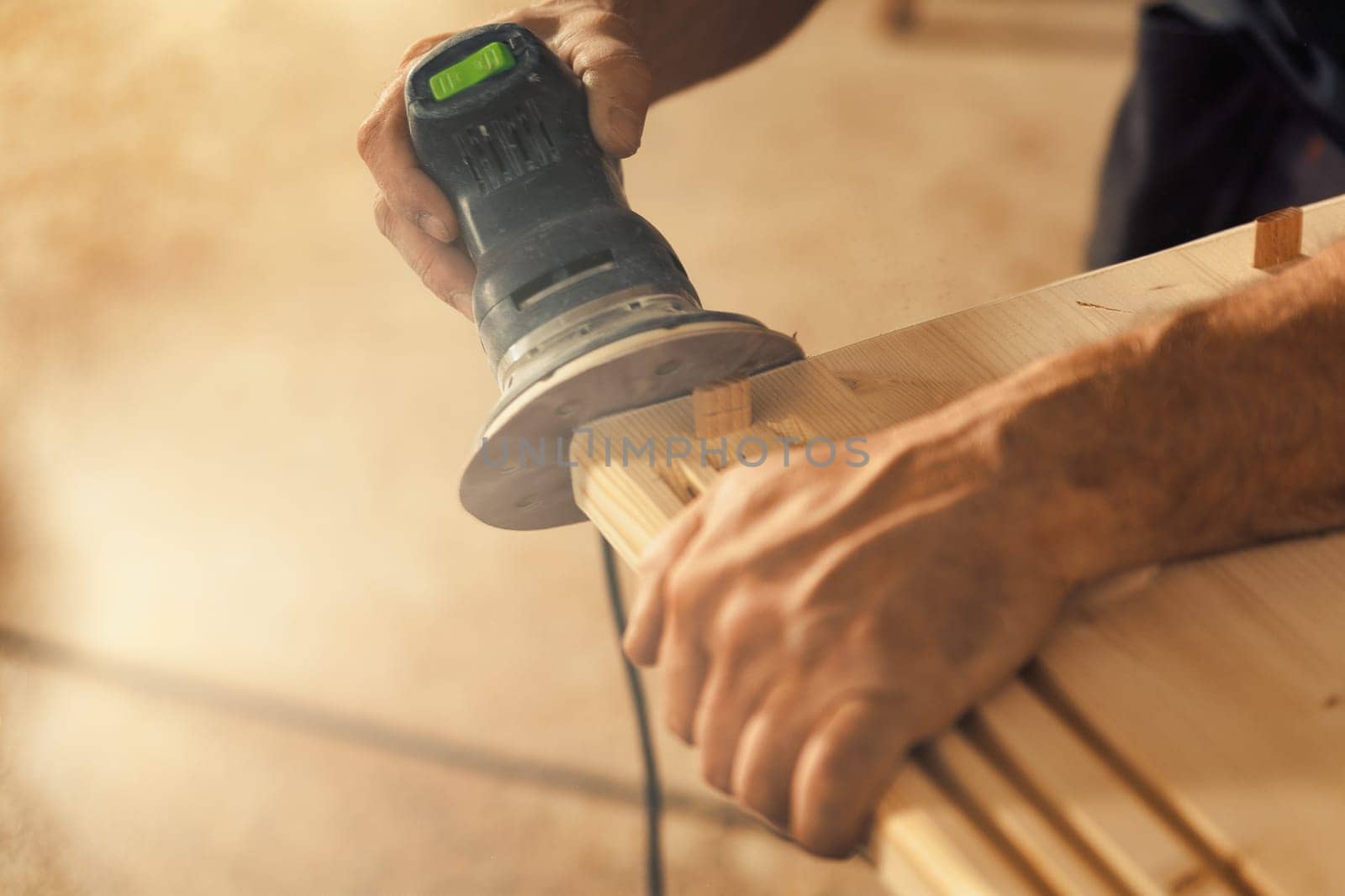 The carpenter's hands grip a sander. The wood artisan, an experienced professional, holds a board or piece of wood with strong and precise hands, carefully sanding it with a machine that vibrates sand
