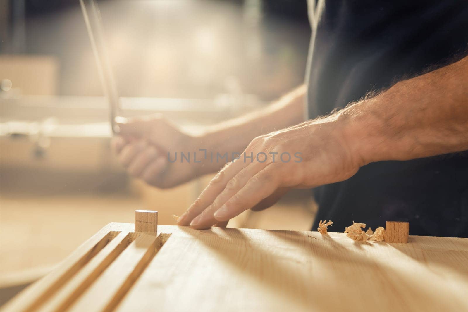 An artisan stands with his drawknife in hand. This carpenter loves to use his manual cutting tool because he desires control and the ability to rely solely on his own hands. In the background, his wor