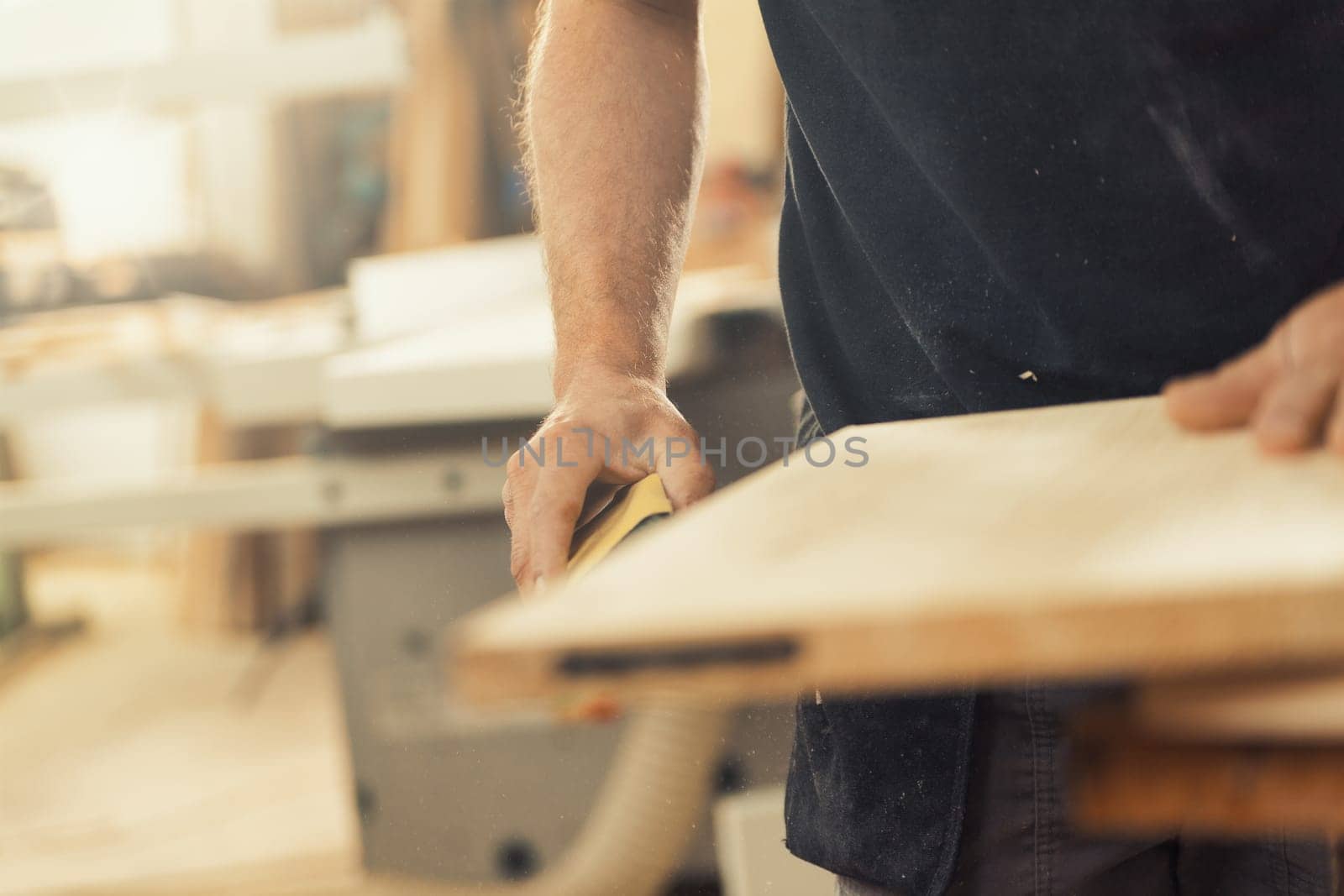 The wood artisan's arm holding a measuring tape is in focus as he prepares to work on the wood in front of him, ready to build furniture. His arm is strong and virile, and the workshop is a mess but a