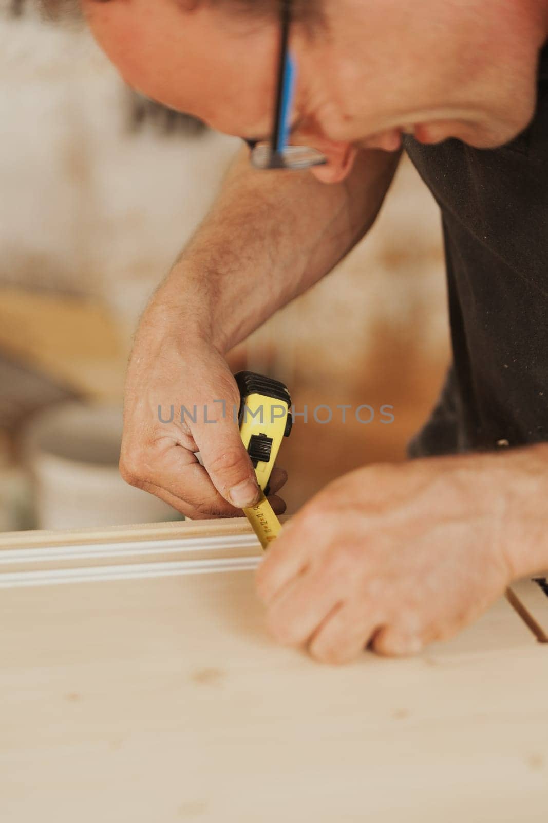 Carpenter squints to get a good read on the measurement he is taking with the pocket tape measure on a component resting on the table. Craftsman builds wooden furniture.