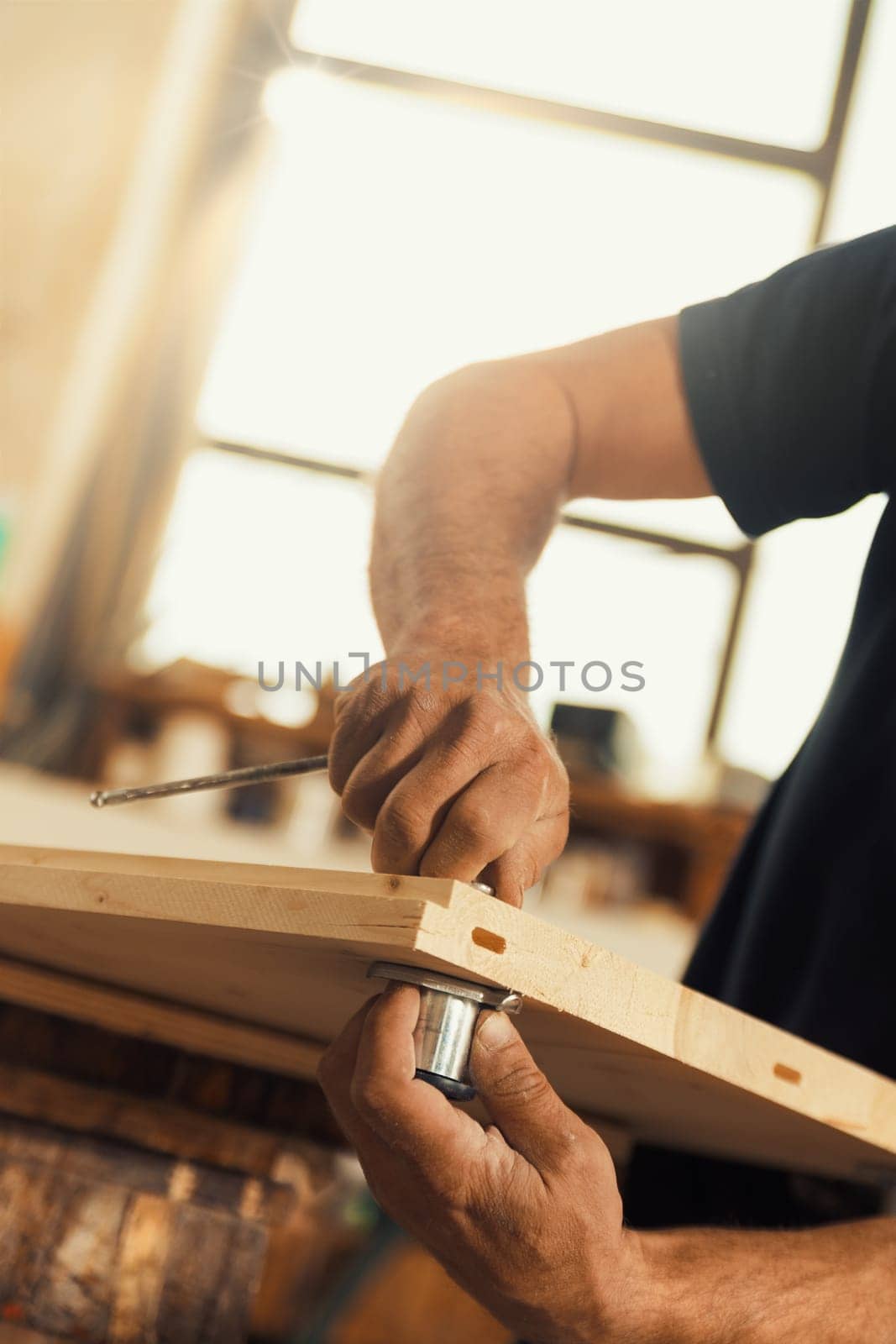 The carpenter screws and secures a wooden board using an Allen key, holding the other side with one hand. Strong, muscular, and hairy arms and hands are visible, exemplifying the manual labor involved