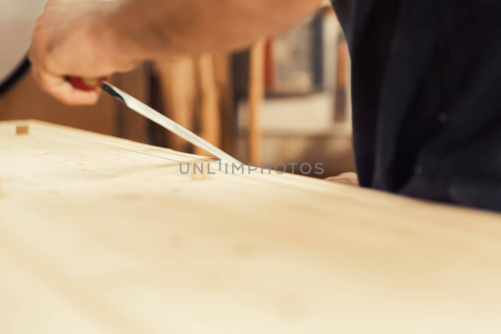 An artisan stands with his drawknife in hand. This carpenter loves to use his manual cutting tool because he desires control and the ability to rely solely on his own hands. In the background, his wor