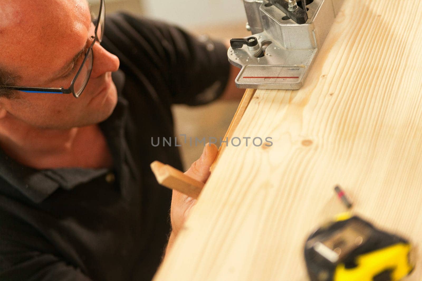 Woodworker focused while using a multi-pattern drillng jig. He wears eyeglasses and there is a measuring tape on the plank he's working on.