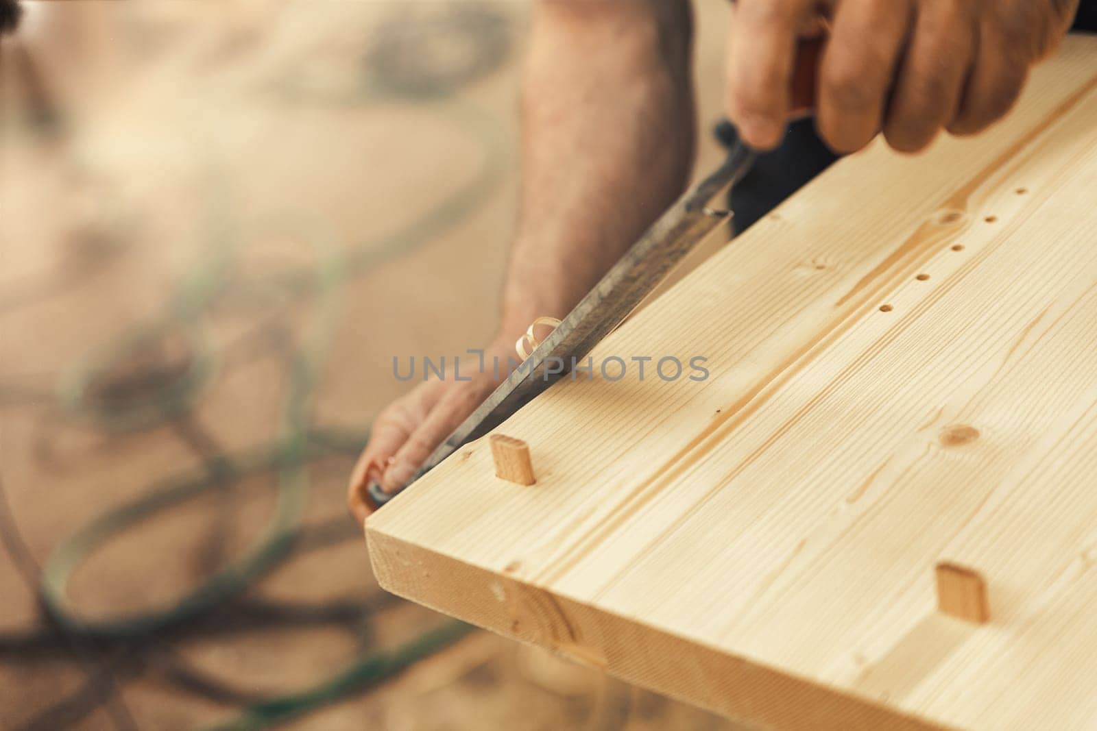 The artisan carves and chamfers the edges of the wooden boards that make up the furniture with a drawknife. Strong male hands and wood are visible, embodying the essence of carpentry