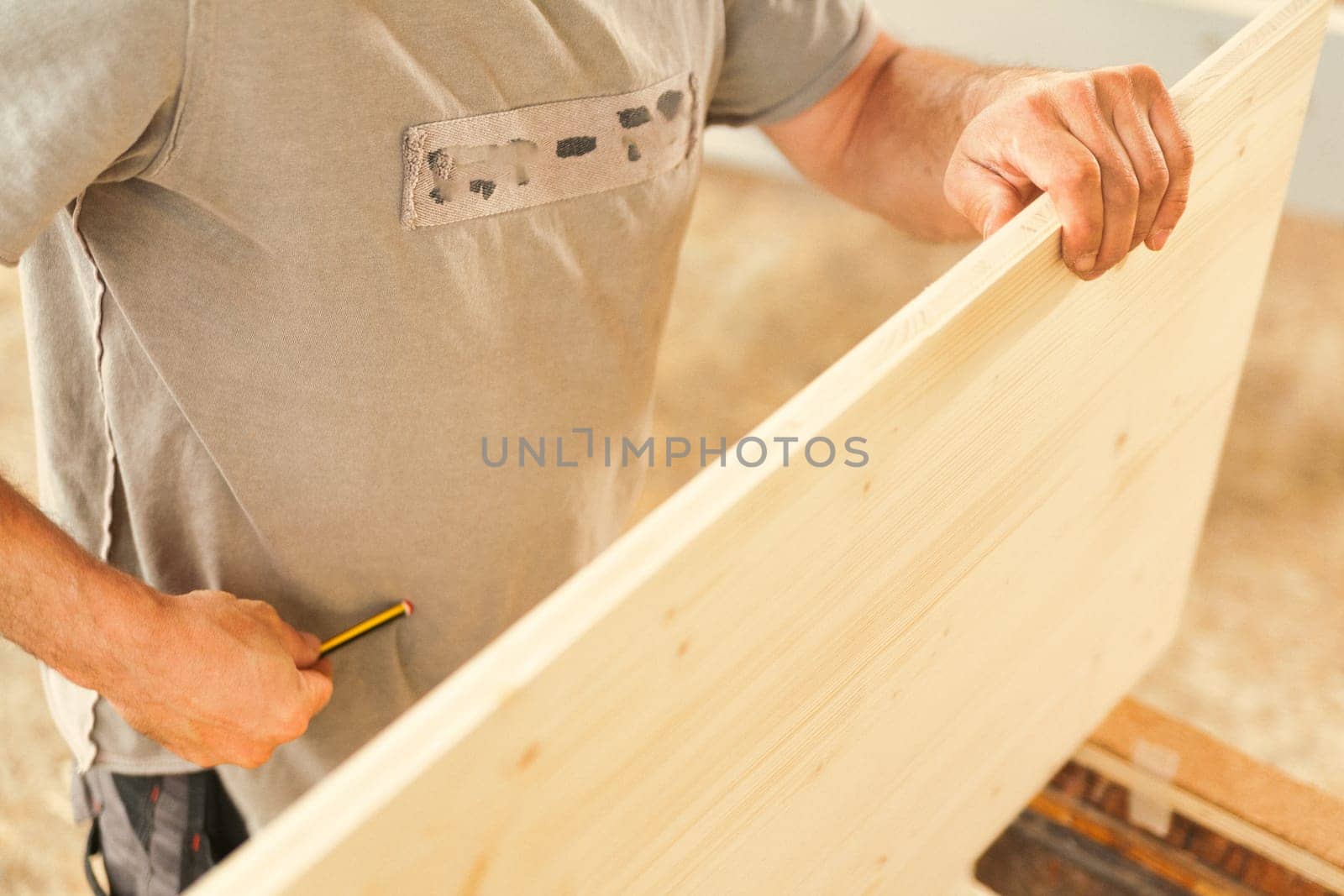 Carpenter holds up a wide rough wooden shelf with one hand as he prepares to use a pencil. Face not visible. Hand in focus and the rest out of focus.
