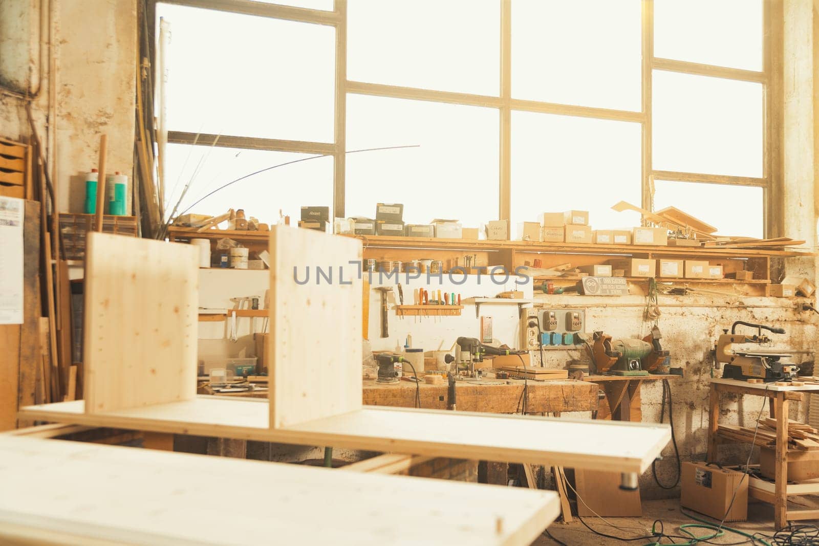 General view of a carpenter's workshop with giant window that illuminates all the creative clutter and pieces of furniture being worked on in the warm sunlight blending into the wood.
