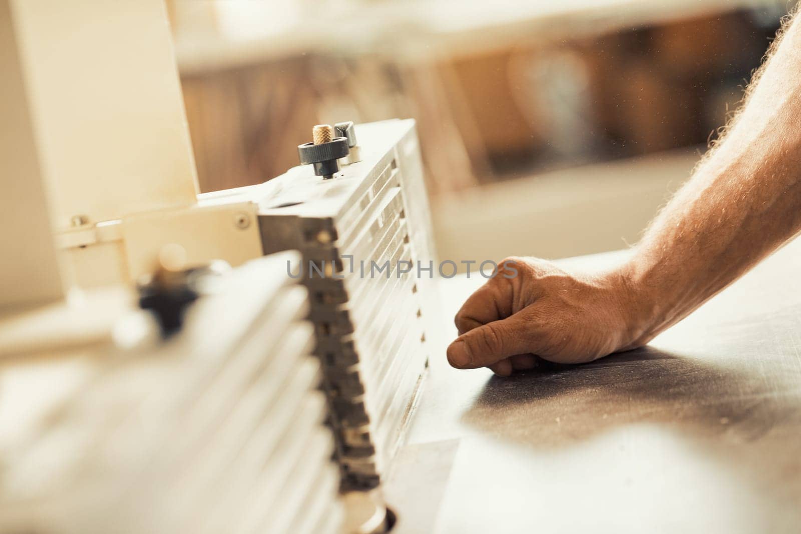 A carpenter's strong and muscular hand and forearm holding a metal tool while working on a self-operating wooden piece of furniture. The warm sunlight flooding the workshop and the wood