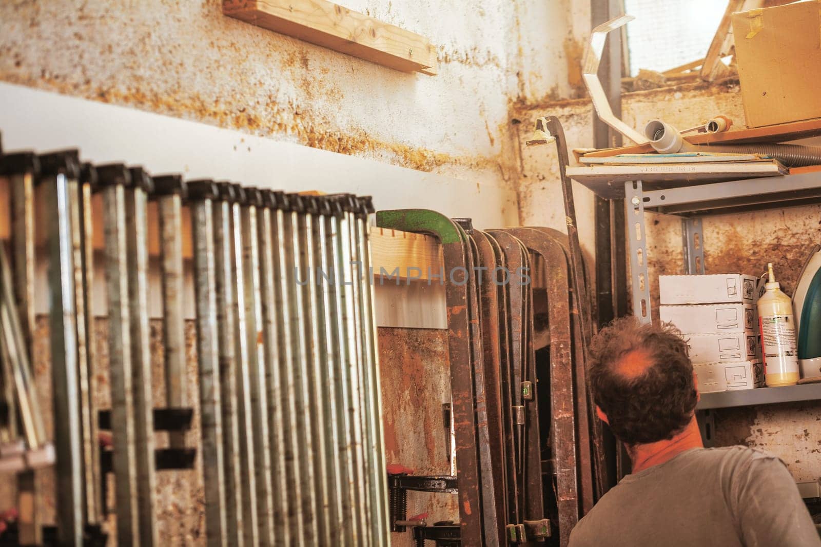 In a traditional craft workshop, lots of wood clamps, large and very large, are lined up on the wall. The carpenter is picking out tools, and sunlight filters through the window.