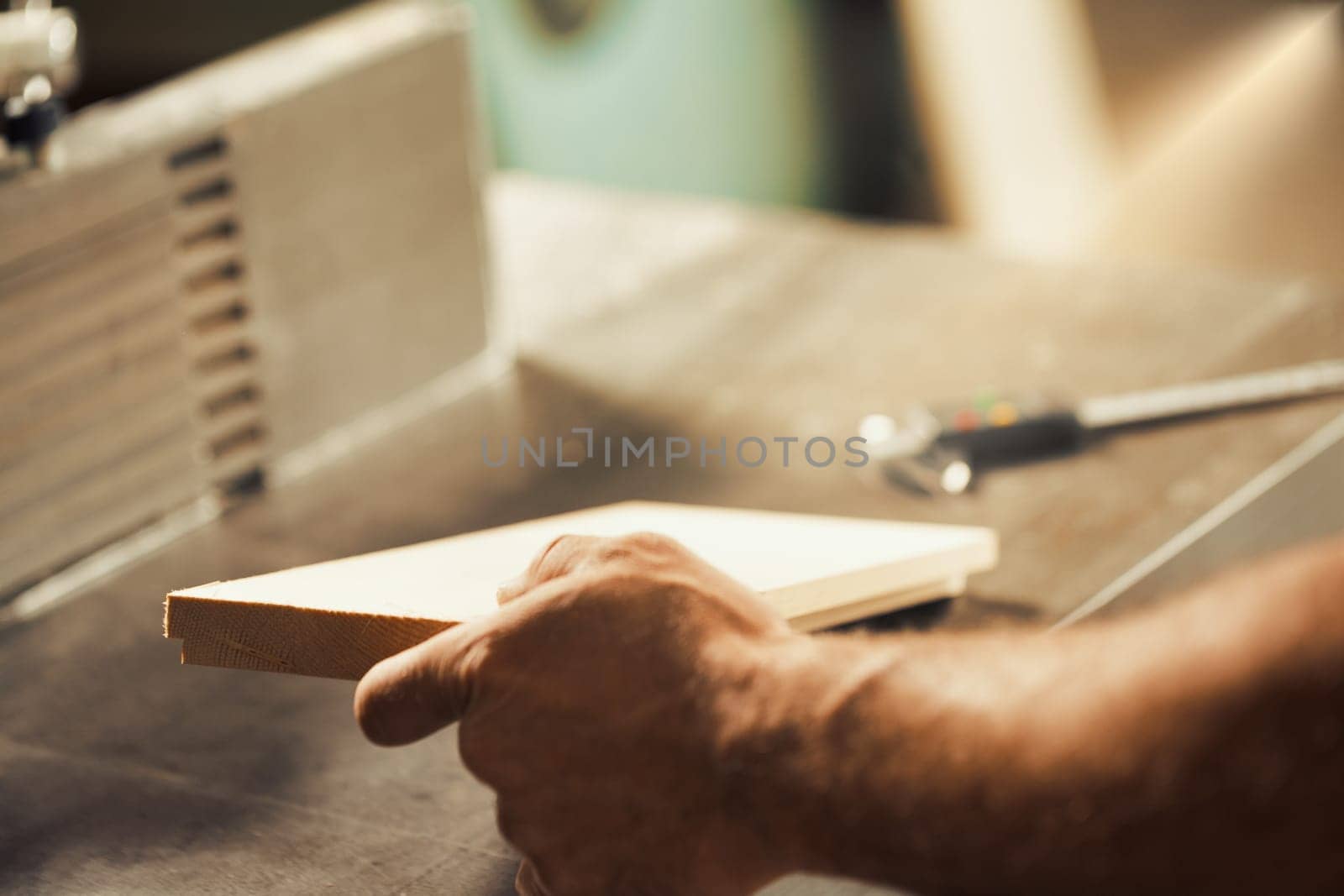 A carpenter's strong and muscular hand and forearm holding a metal tool while working on a self-operating wooden piece of furniture. The warm sunlight flooding the workshop and the wood