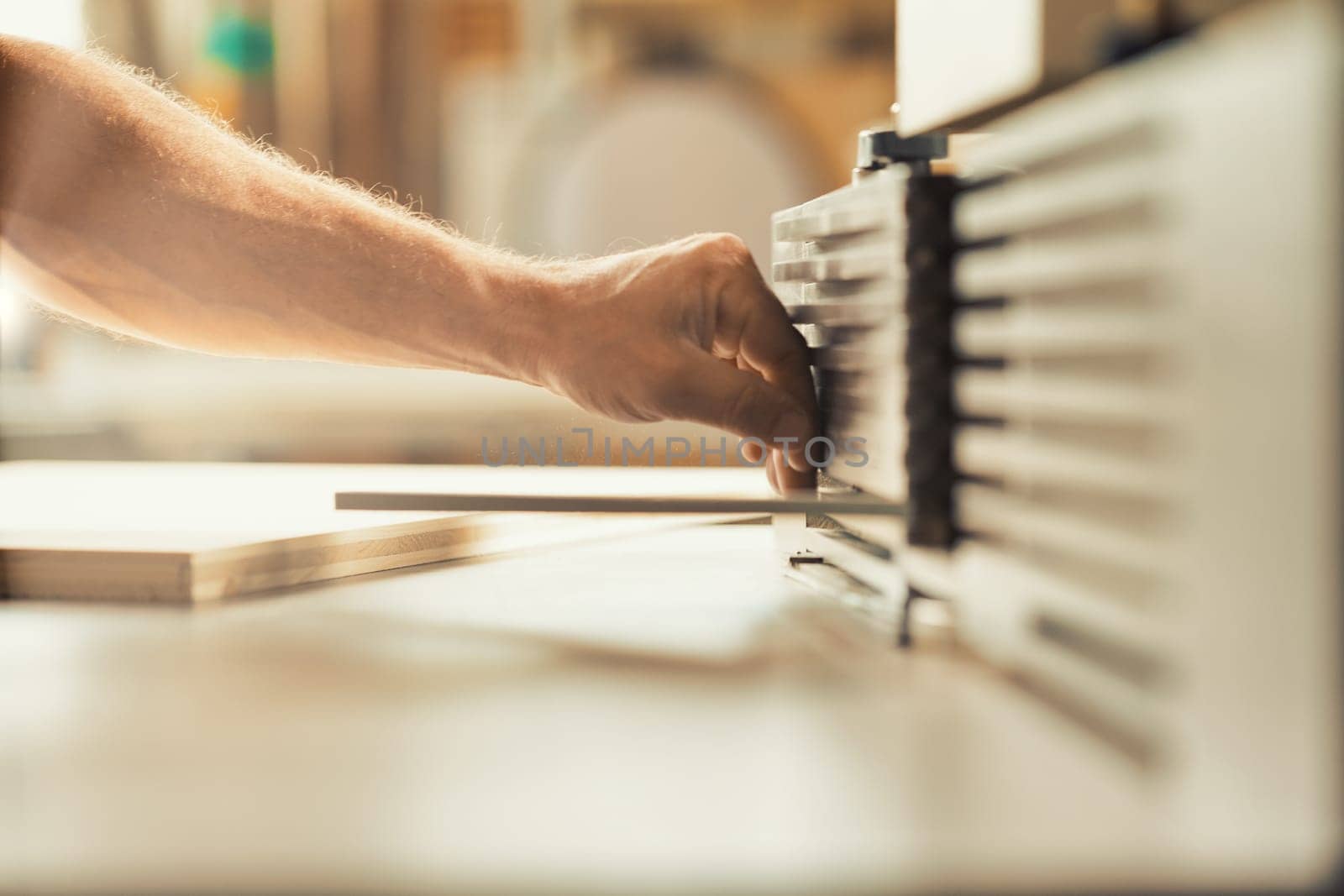 A carpenter's strong and muscular hand and forearm holding a metal tool while working on a self-operating wooden piece of furniture. The warm sunlight flooding the workshop and the wood