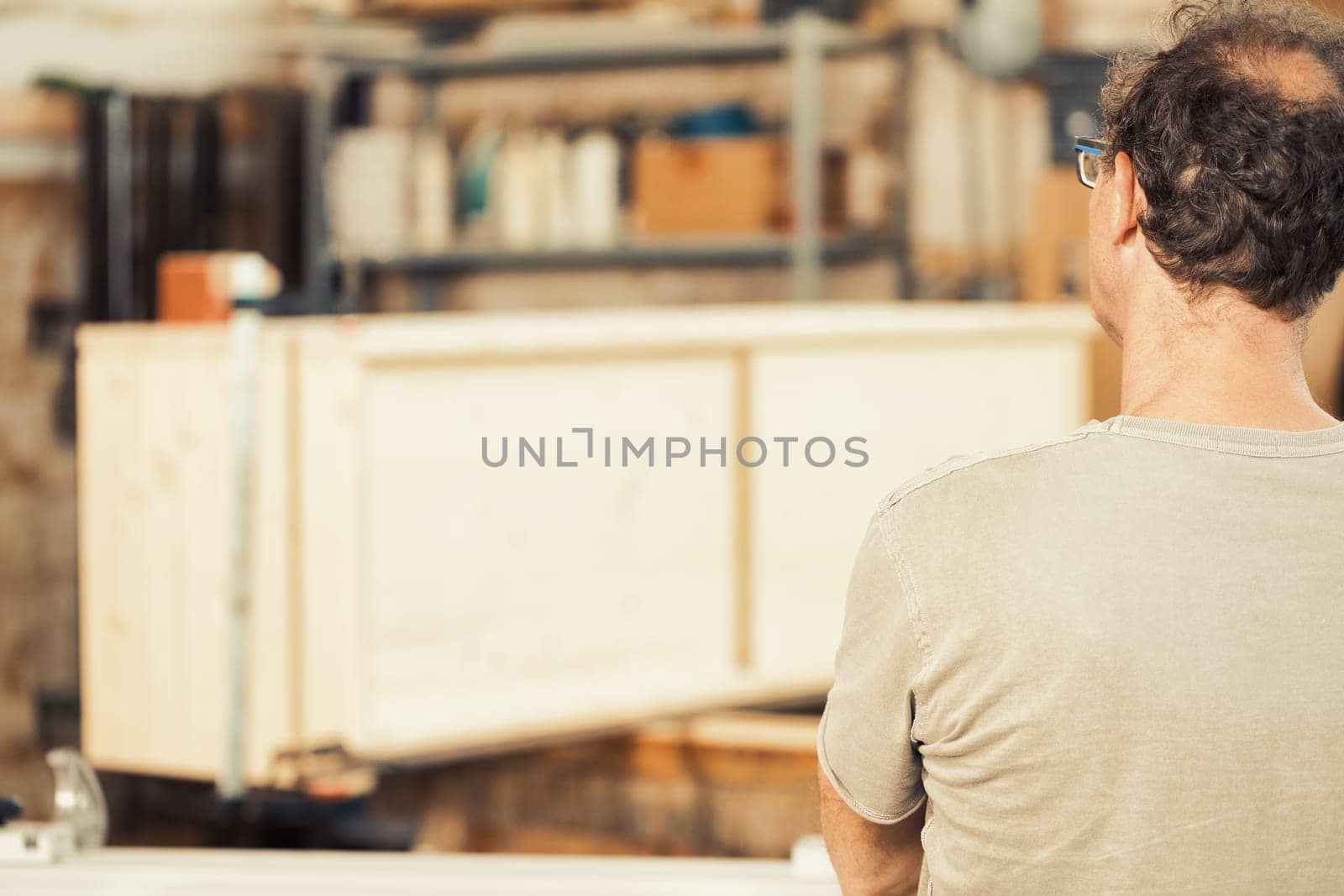 Carpenter examines furniture he's working on to determine the best approach. He's in the foreground, in focus, while the background shows the out-of-focus work and controlled chaos of his workshop