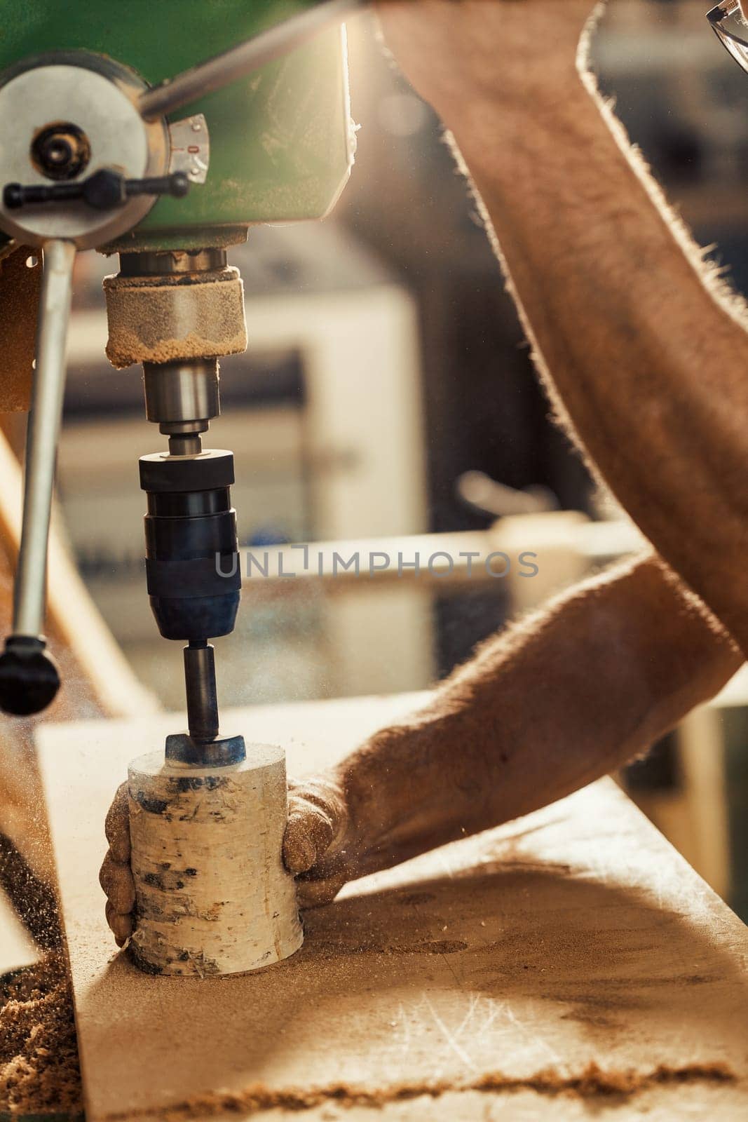 The carpenter's strong hand firmly holds a birch log while using a drill press to carve it, lowering the lever with his other arm. The artisanal workshop is backlit, and sawdust and wood chips can be