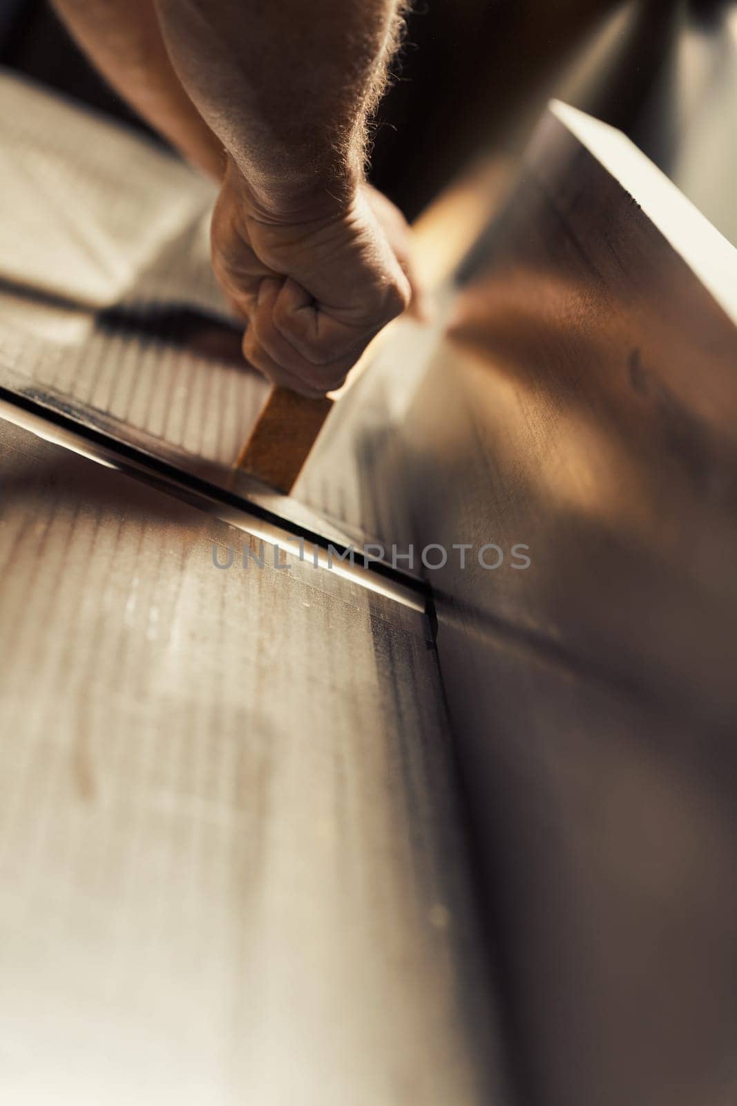 The strong and muscular hand and forearm of a carpenter holding a metal tool, using it to aid in the construction of a piece of furniture that will be entirely crafted by him. The warm sunlight floodi