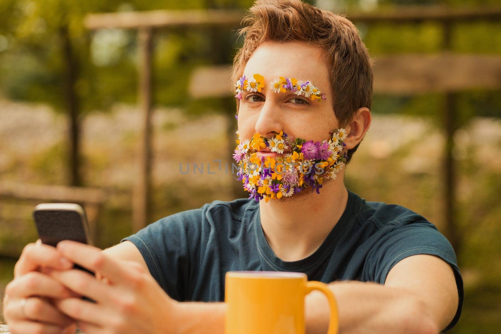 Young man with colorful spring flowers instead of facial hair sits amidst nature. While cherishing the tranquility and fresh air, he doesn't despise technology, staying connected via phone