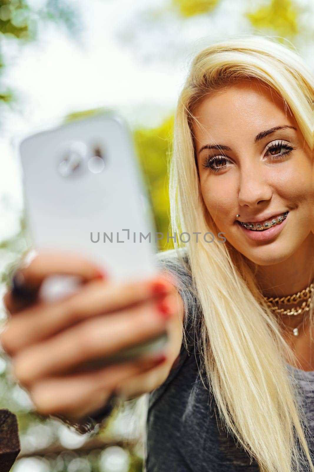 Beautiful blond girl stares amusedly at her own cell phone. Her smile leaves her teeth uncovered with braces which still makes her super cute. she is outdoors in nature.