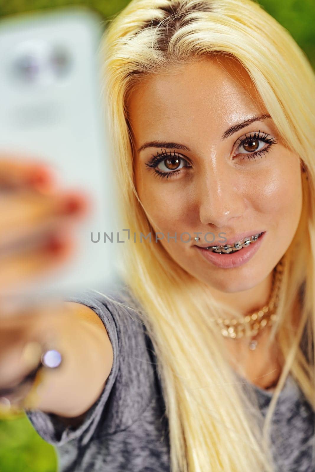 Vertical portrait of young blond woman in selfie position. Fixes the lens with intense eye-contact. She smiles highlighting her orthodontic braces that nonetheless make her even more attractive. Compl