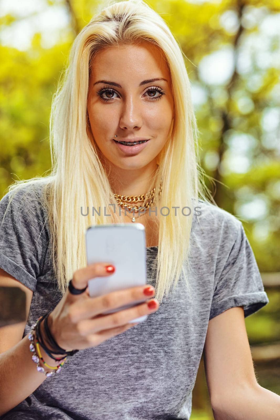 Beautiful blond girl in an outdoor forest or public park on a sunny day. She has very visible braces that make her even prettier. She has a smartphone that she has stopped looking at to turn her atten