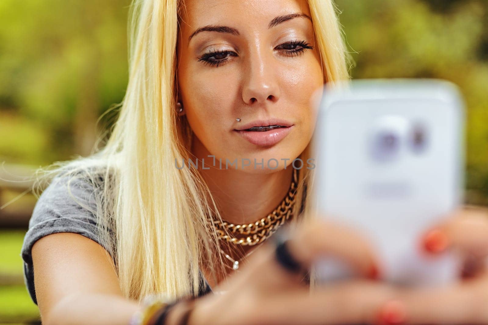 Beautiful blond girl in an outdoor forest or public park on a sunny day. She has very visible braces that make her even prettier. She is looking at something in a smartphone screen in a blurred foregr