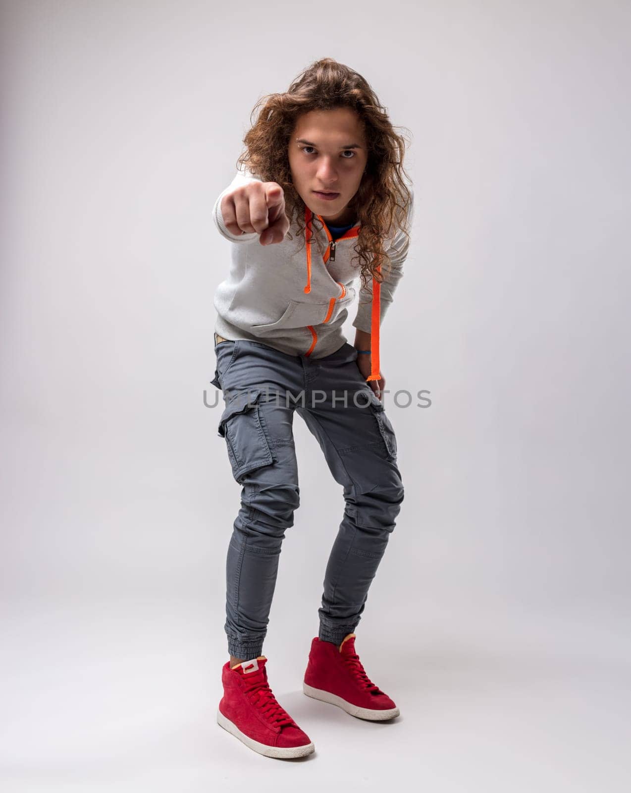 Portrait of young man with long hair. He is wearing ankle-high skinny jeans and high-top sneakers and a gray sweatshirt with neon orange details. he is standing but slightly crouched, pointing at us i