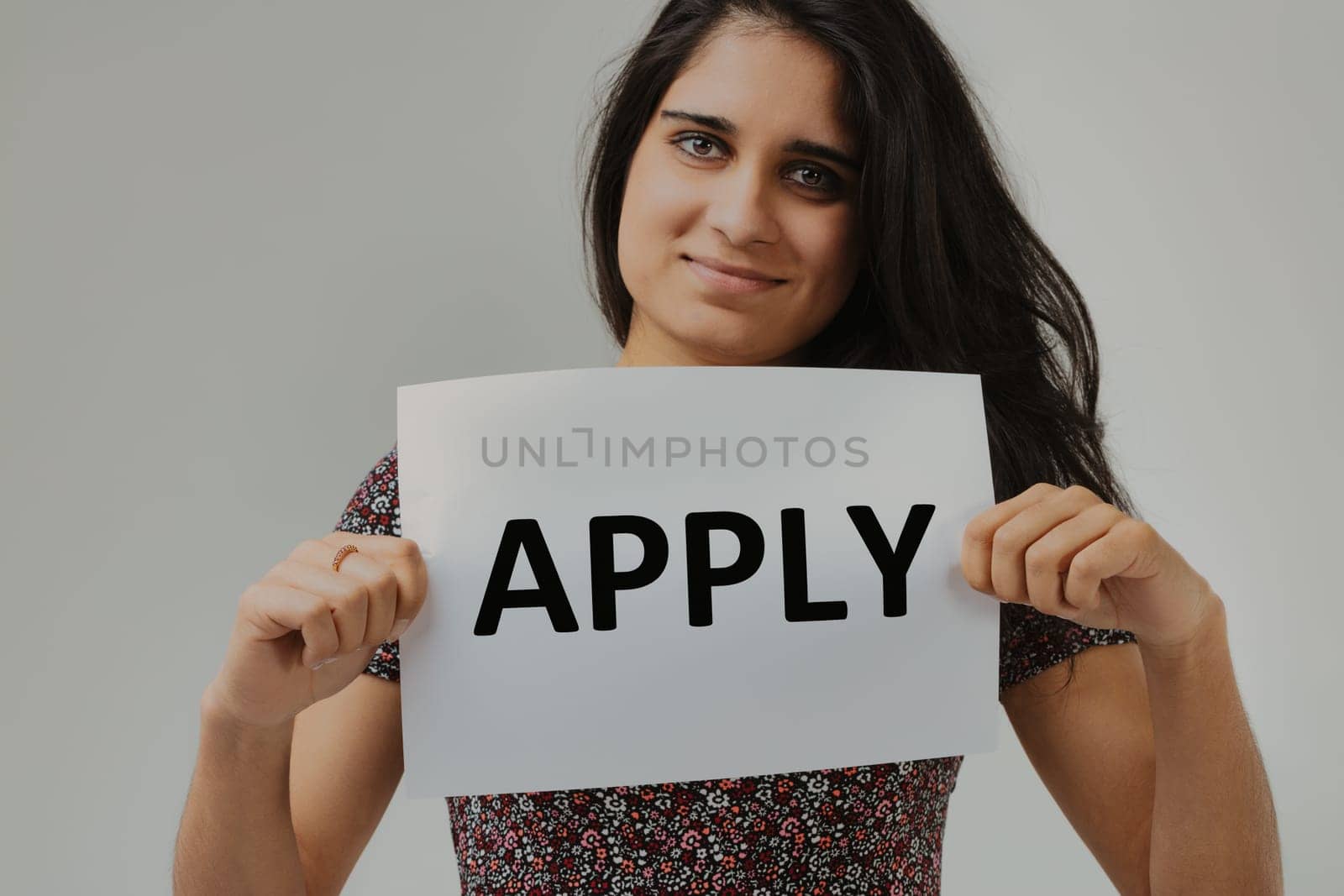 With a sign that simply reads 'APPLY,' a woman with striking eyes and a welcoming demeanor stands as the messenger. Her outfit's design accentuates the overall composition