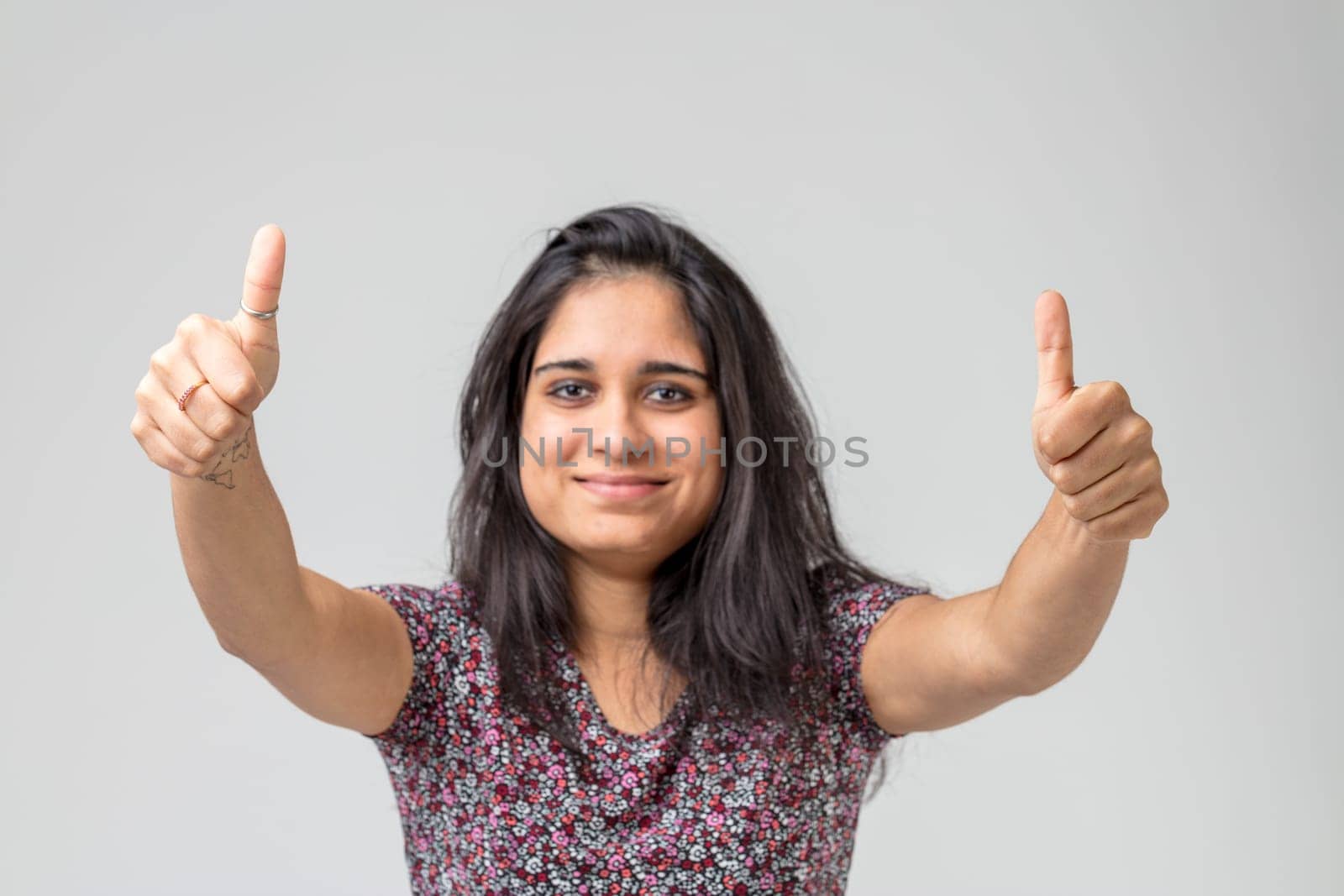 Thumbs up for you from a young Indian woman with long black hair, smiling and wearing a flowered dress. Her hands are in focus while she, behind, is out of focus. Concept of okay, positivity and movin