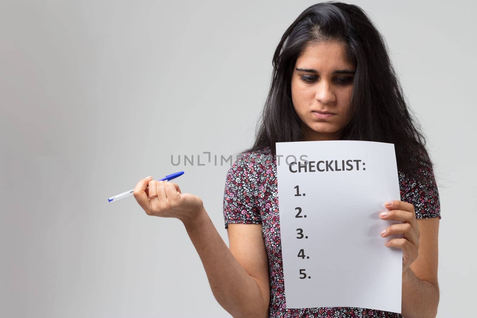 Young Indian woman or girl with long black hair and a flowery dress who could be as much a student as a novice worker. She holds a checklist in one hand, which she stares at analyzing it, and a pen in