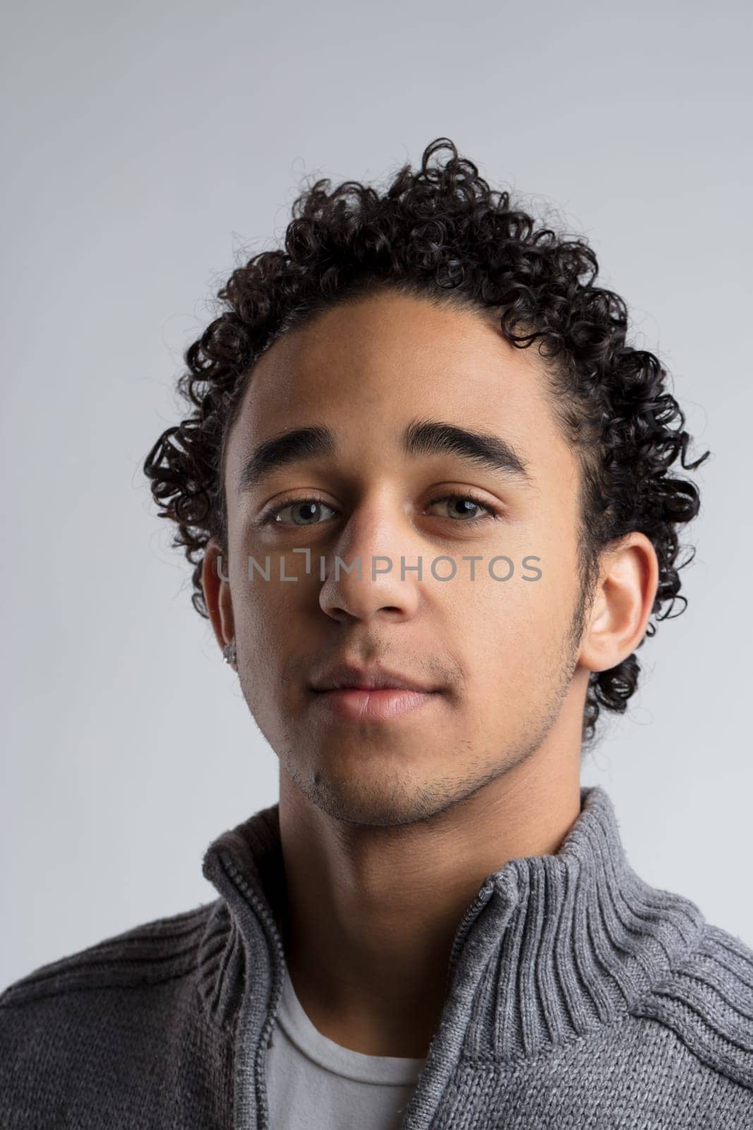 Captured against a neutral backdrop, a young man with curly dark hair exudes a sense of contemplation, his green eyes hinting at a story