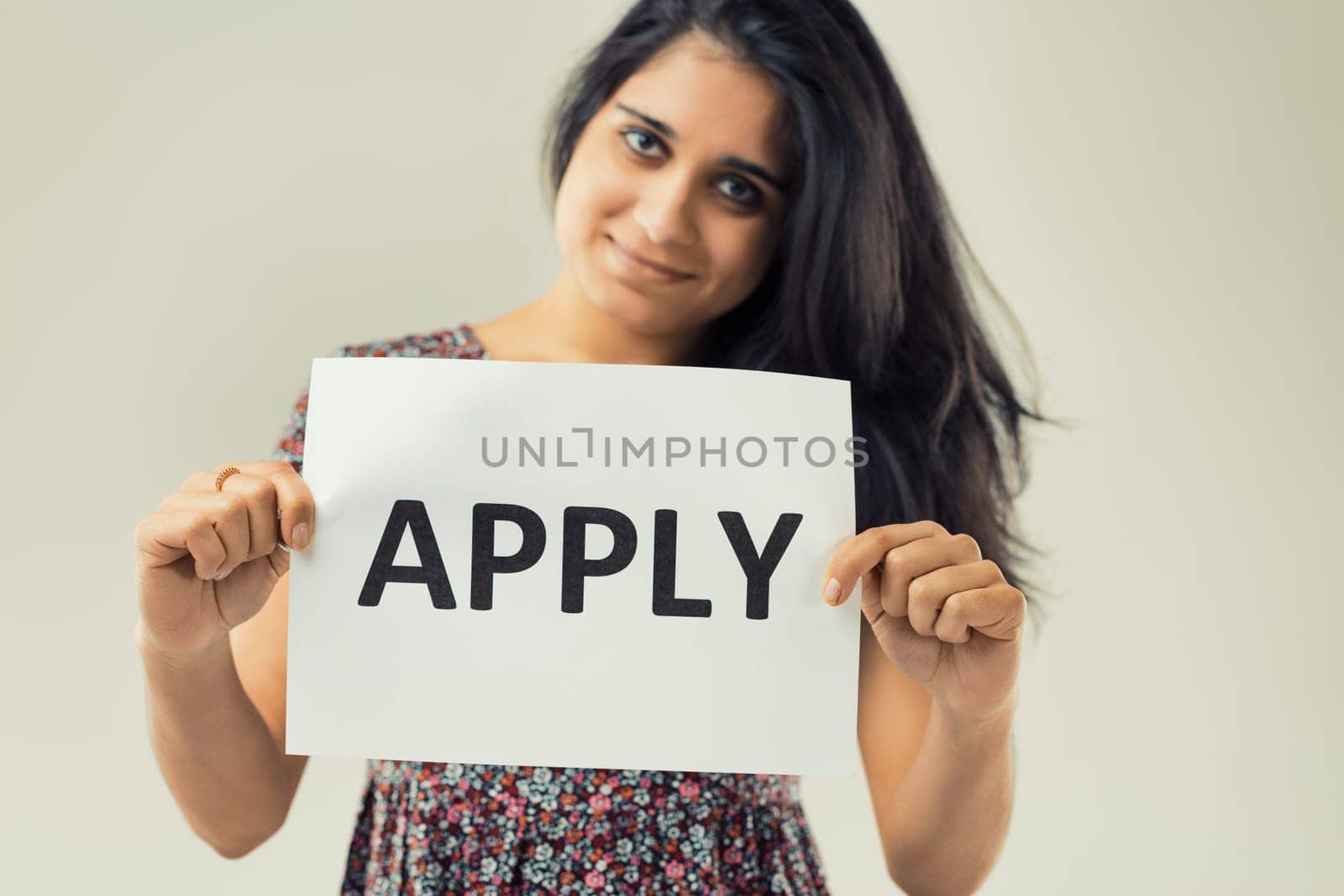 woman with expressive eyes and flowing dark hair confidently holds a sign stating 'APPLY.' The floral pattern of her attire contrasts the simple, clear message she's conveying