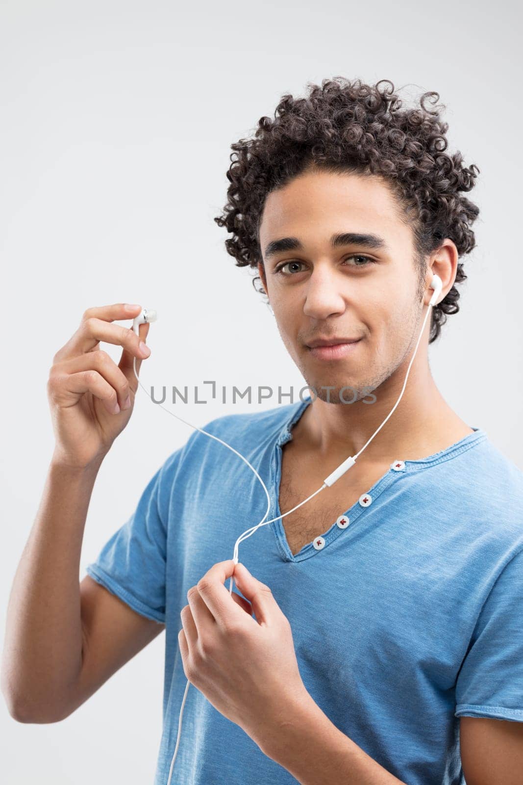 Young smiling man with curly hair showcases white earbuds, wearing a blue shirt