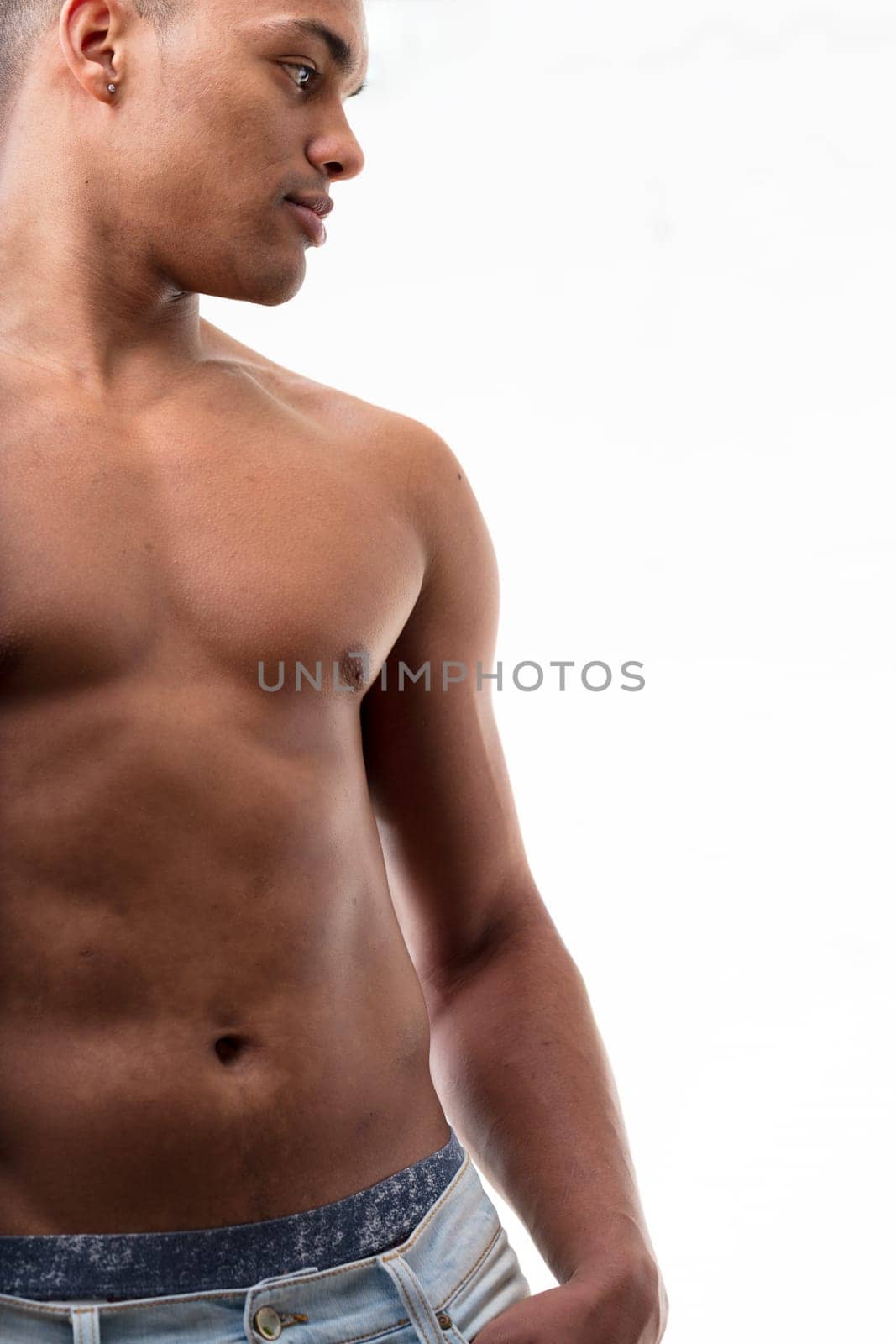 A fit young man, shirtless, gazes sideways. His undergarments visible beneath his trousers, his thumb casually hooked into a pocket. His chest and abs in view, skin glistening, isolated from the backg