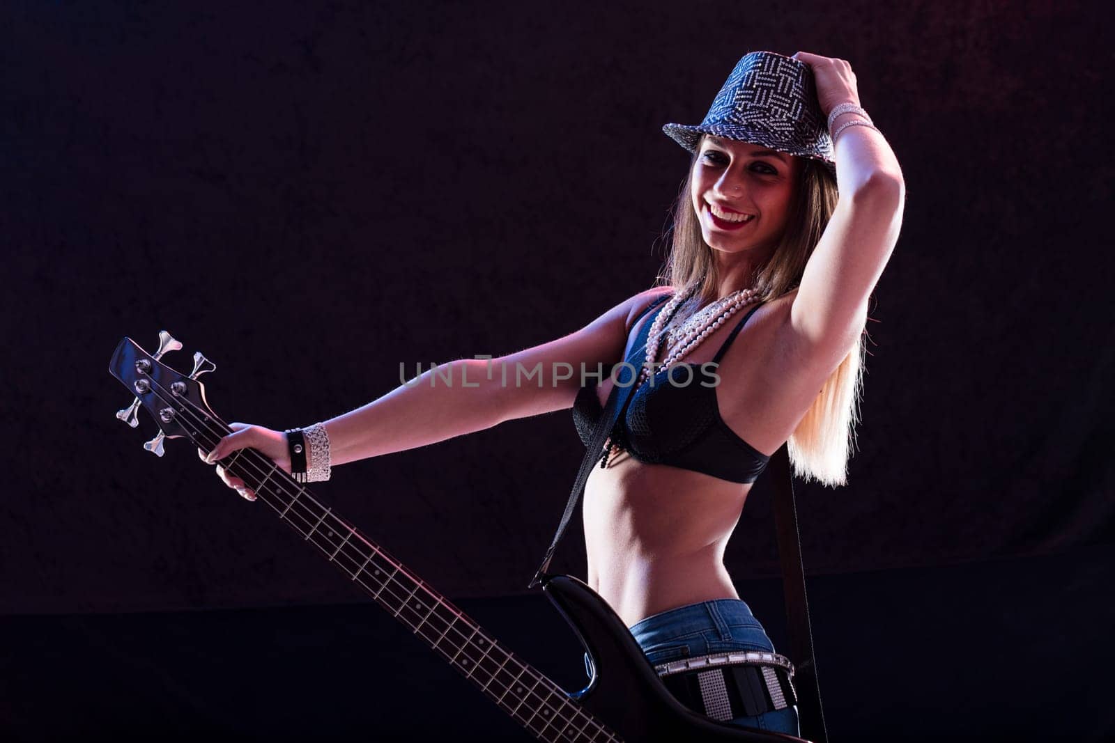 Sexy pop-rock star playing bass guitar with energy. Dressed in high heels, skinny jeans, black bra, and glimmering belts, she moves with confidence, showcasing her toned abs