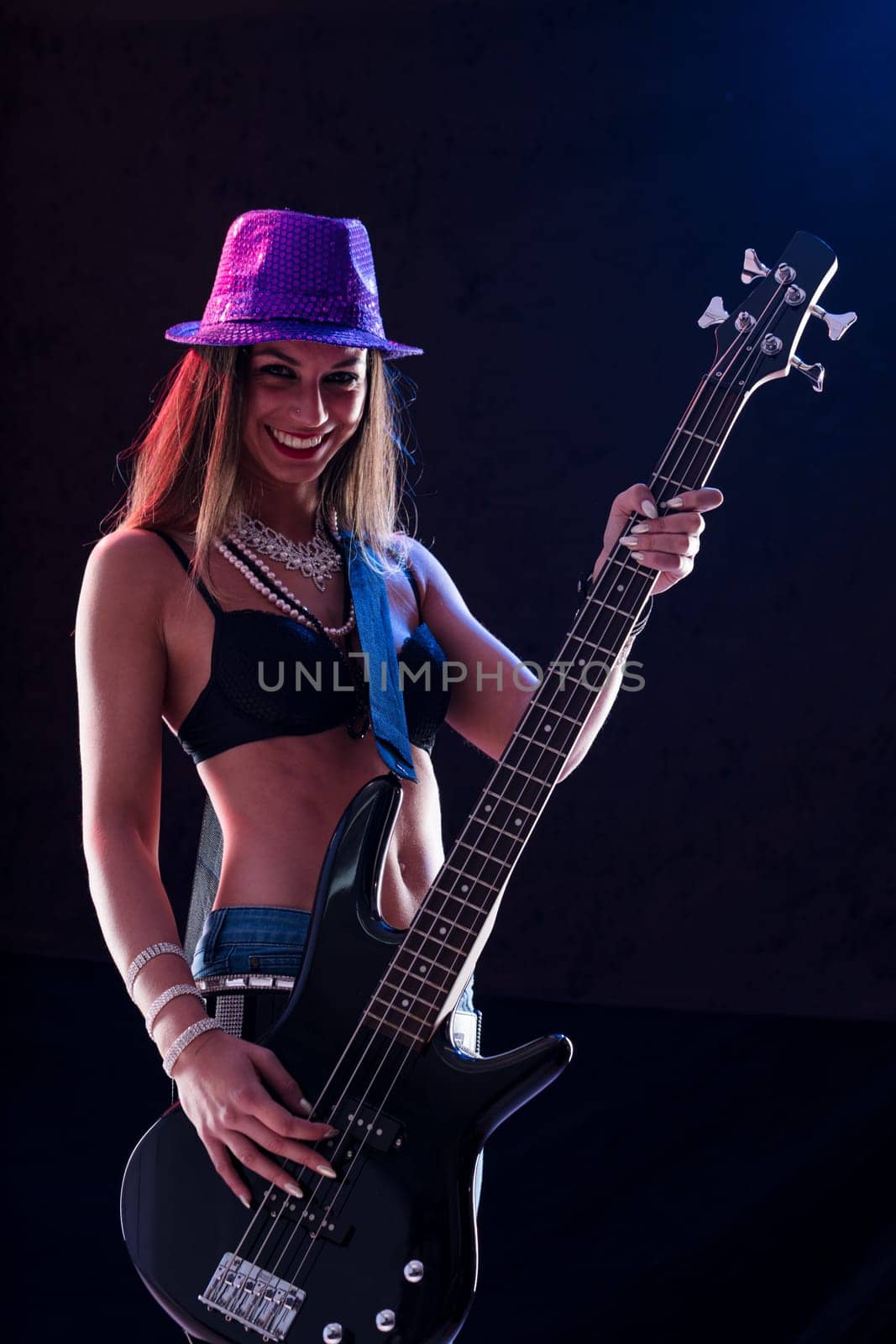 An energetic pop-rock star strums a bass guitar. She's dressed in high heels, skinny jeans, a black bra, and jewel belts, exuding confidence on stage with her flat, muscular abdomen