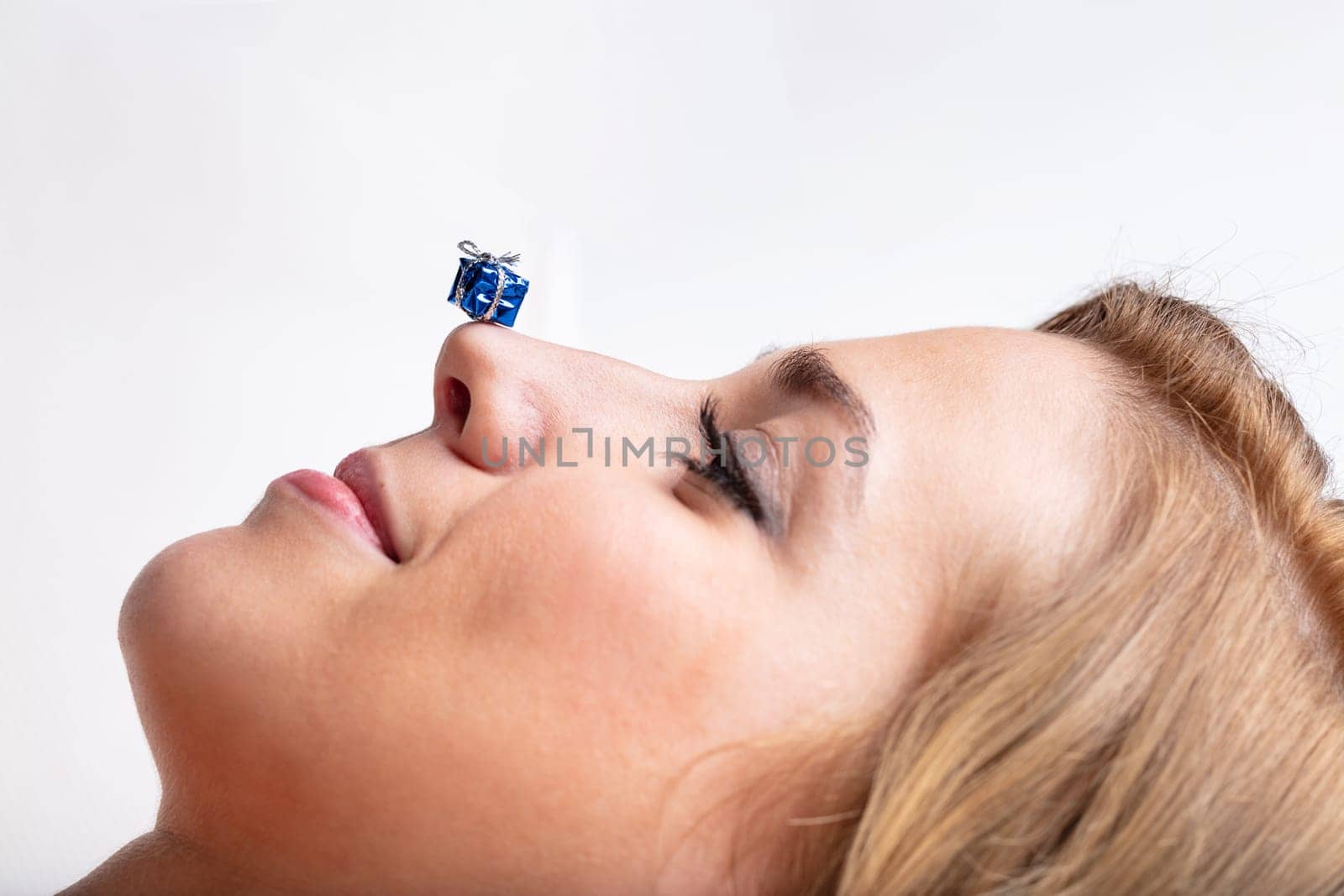 Profile portrait of a slightly surprised and smiling young woman, with a tiny gift resting on her nose tip. Concept of gifts, Christmas, holidays, celebrations  white background