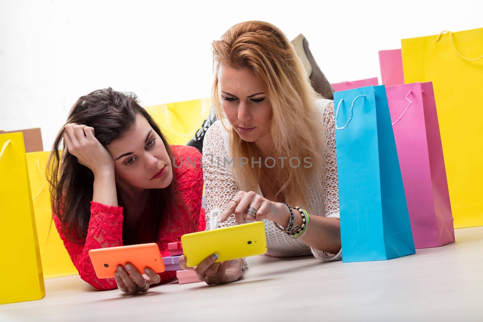 Two women enjoy a break after shopping, browsing on their phones amidst colorful bags