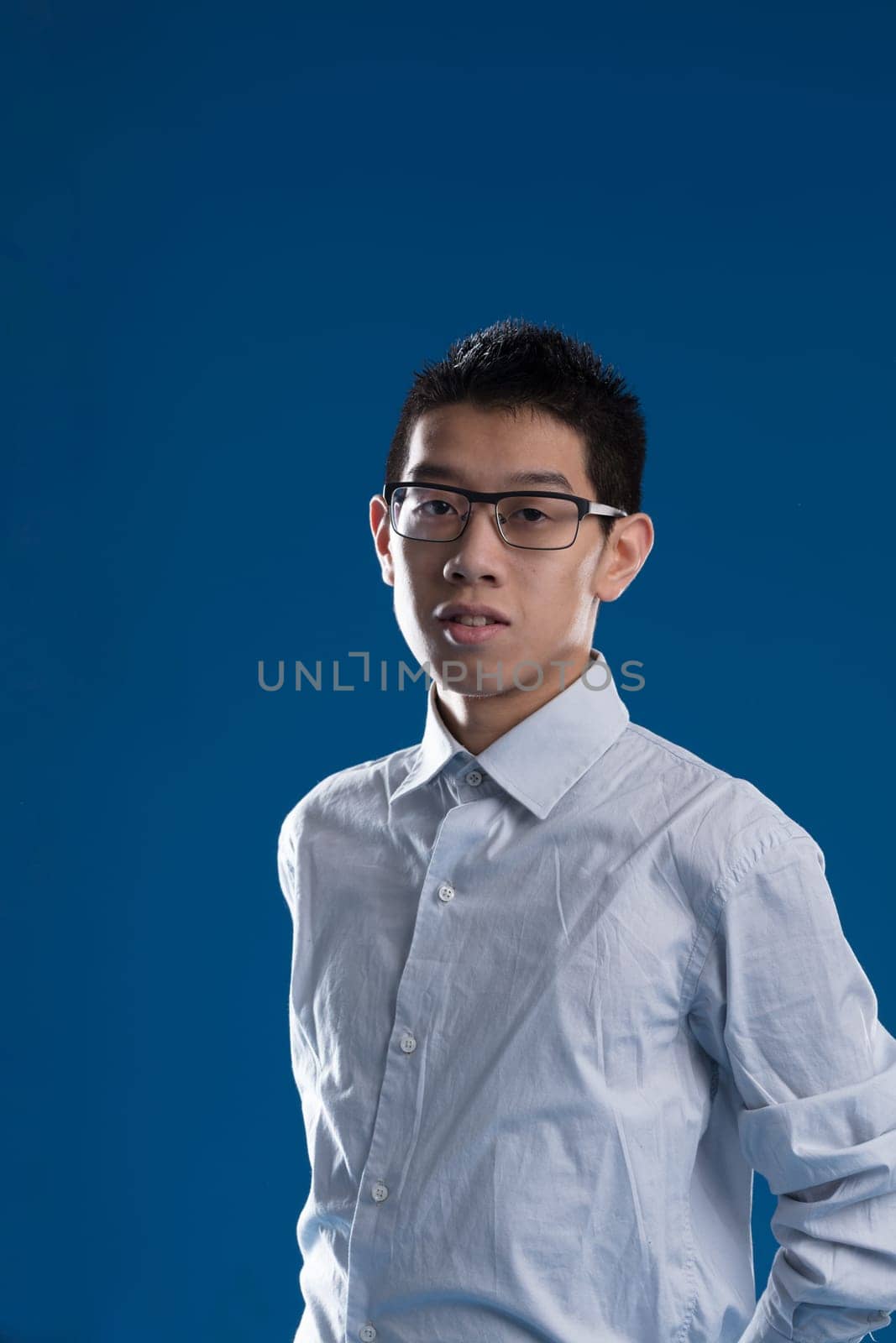 In a half-bust portrait, a young East Asian man wearing glasses and a shirt appears serious and somewhat perplexed. A generous amount of blue copyspace frames him
