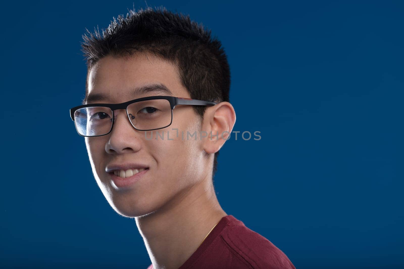 Portrait of a young East Asian man wearing glasses and smiling in a burgundy T-shirt. Short-haired, against a blue background, his image radiates future success and positivity
