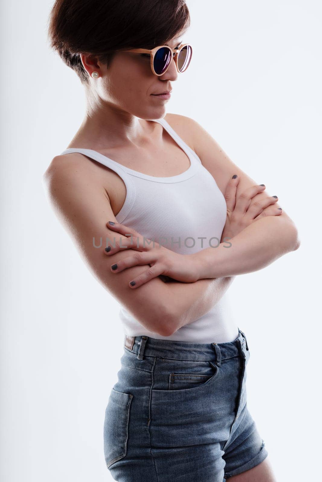 Sexy young woman in trendy shades or sunglasses in a cropped side view on white with folded arms in denim shorts