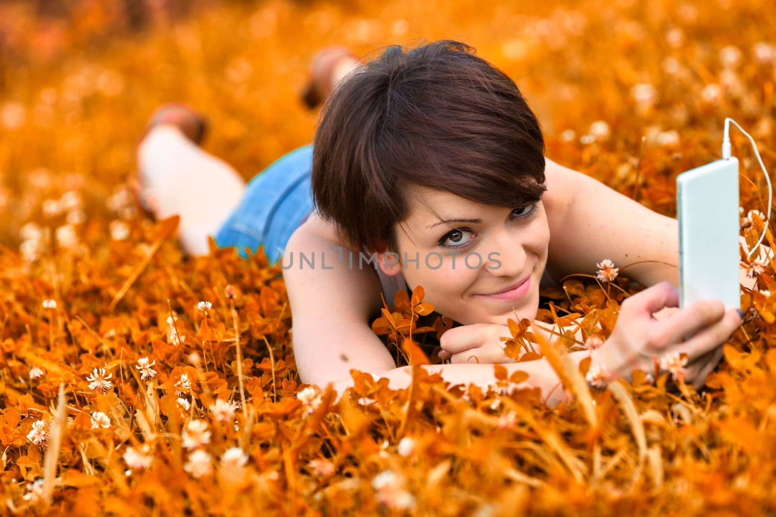 Cute attractive young woman lying in a meadow of orange clover listening to music on her mobile phone in a head to toe view as she smiles at the camera