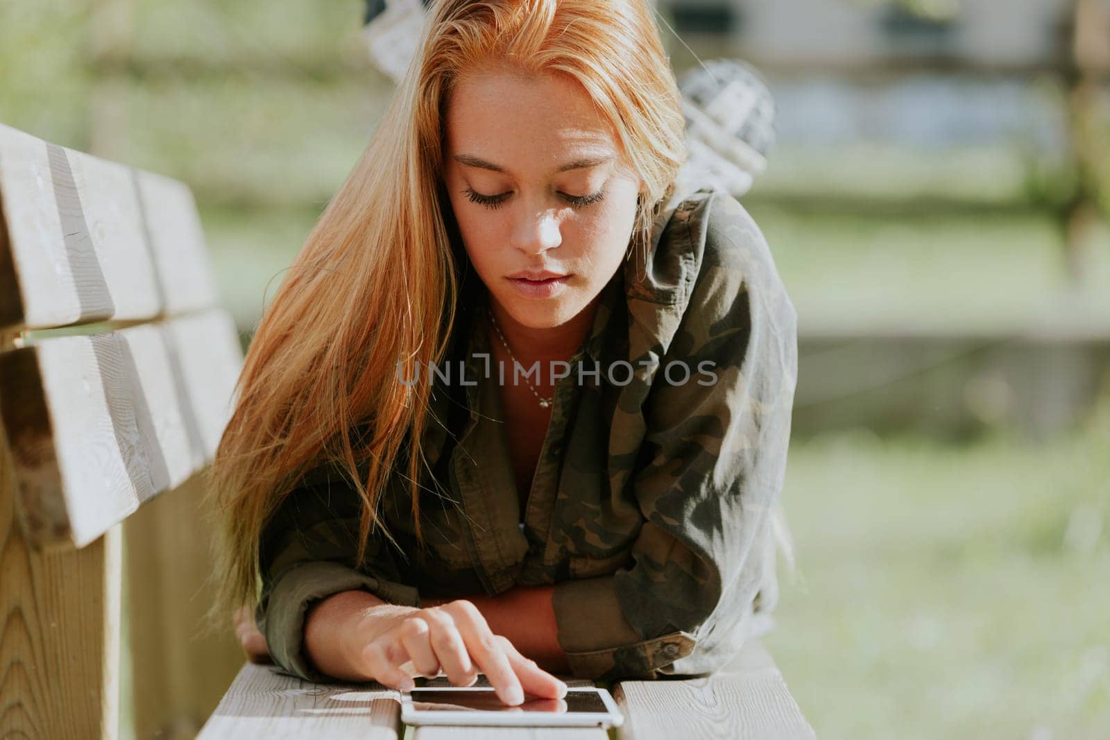 Beautiful blonde girl in casual-sporty attire fiddles with a digital tablet while lying on a park bench, using the internet, studying, working, or having fun