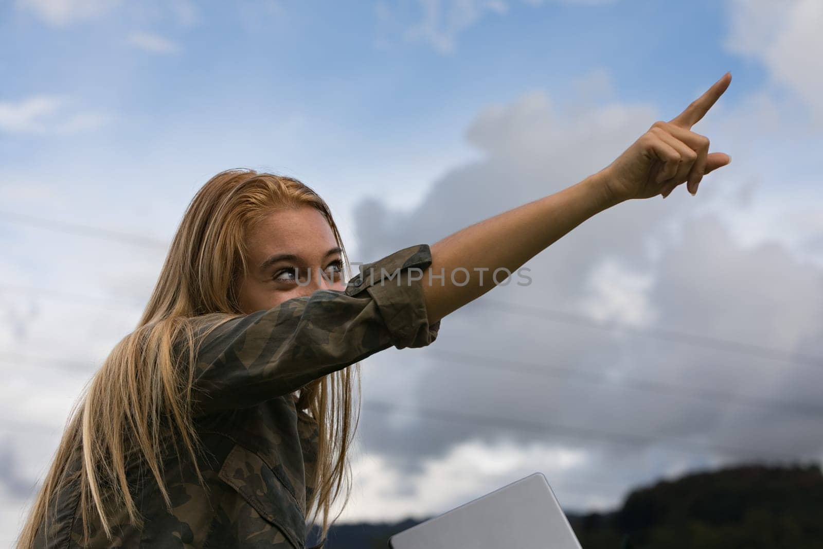 gaze of an androgynous girl in a young blond woman points to the sky with her index finger, but behind her are high voltage cables carrying electricity she has a tablet in her hand