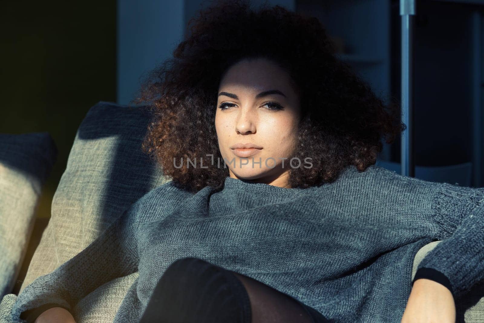 A fiercely confident young woman, successful and prosperous, lounges on her sofa in her spacious living room. Her curly hair, grey sweater, and beauty reflect her talent and dedication